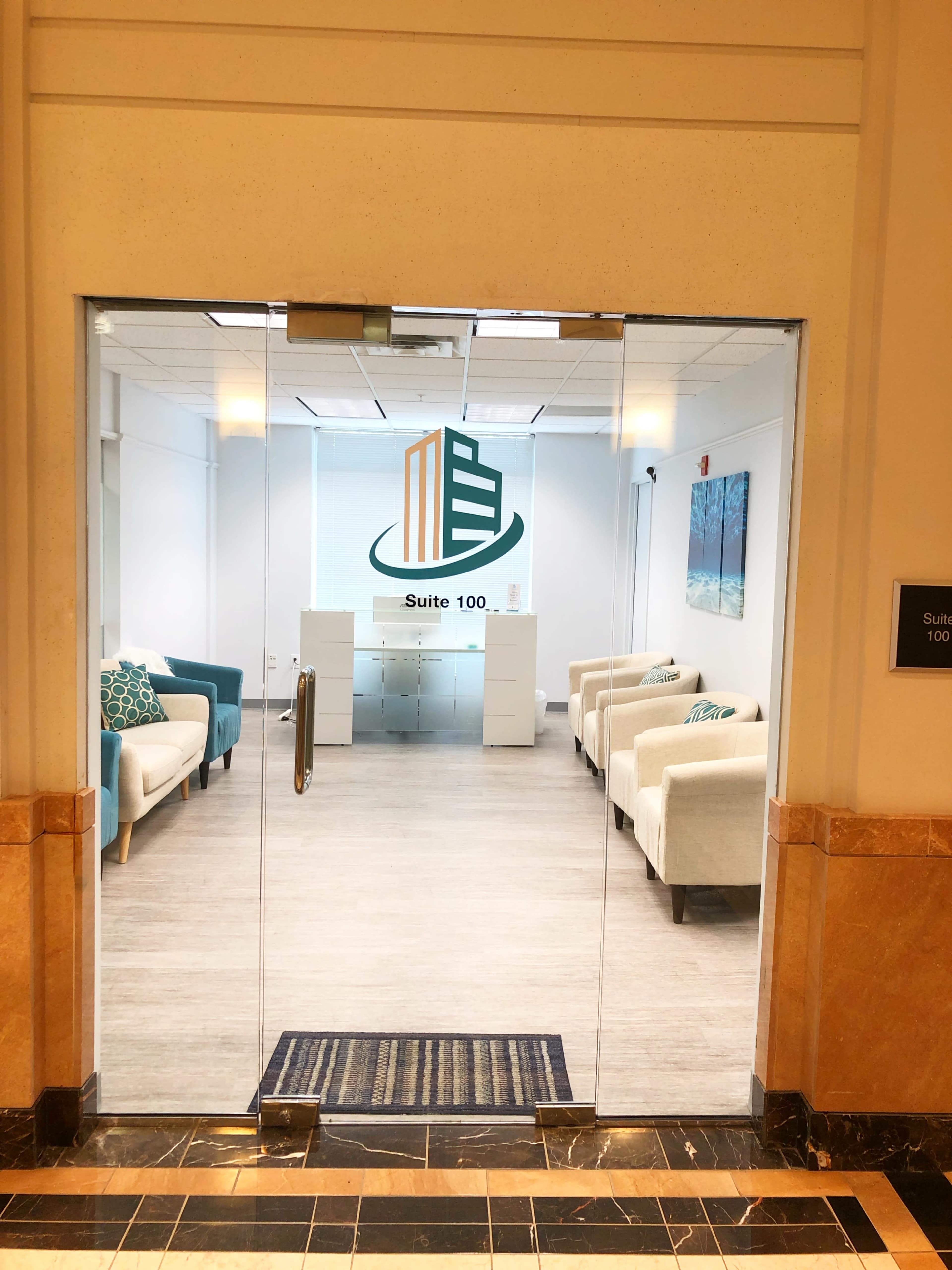 A clear glass door leading into a spacious waiting area with light-colored seating and a reception desk in Suite 100.