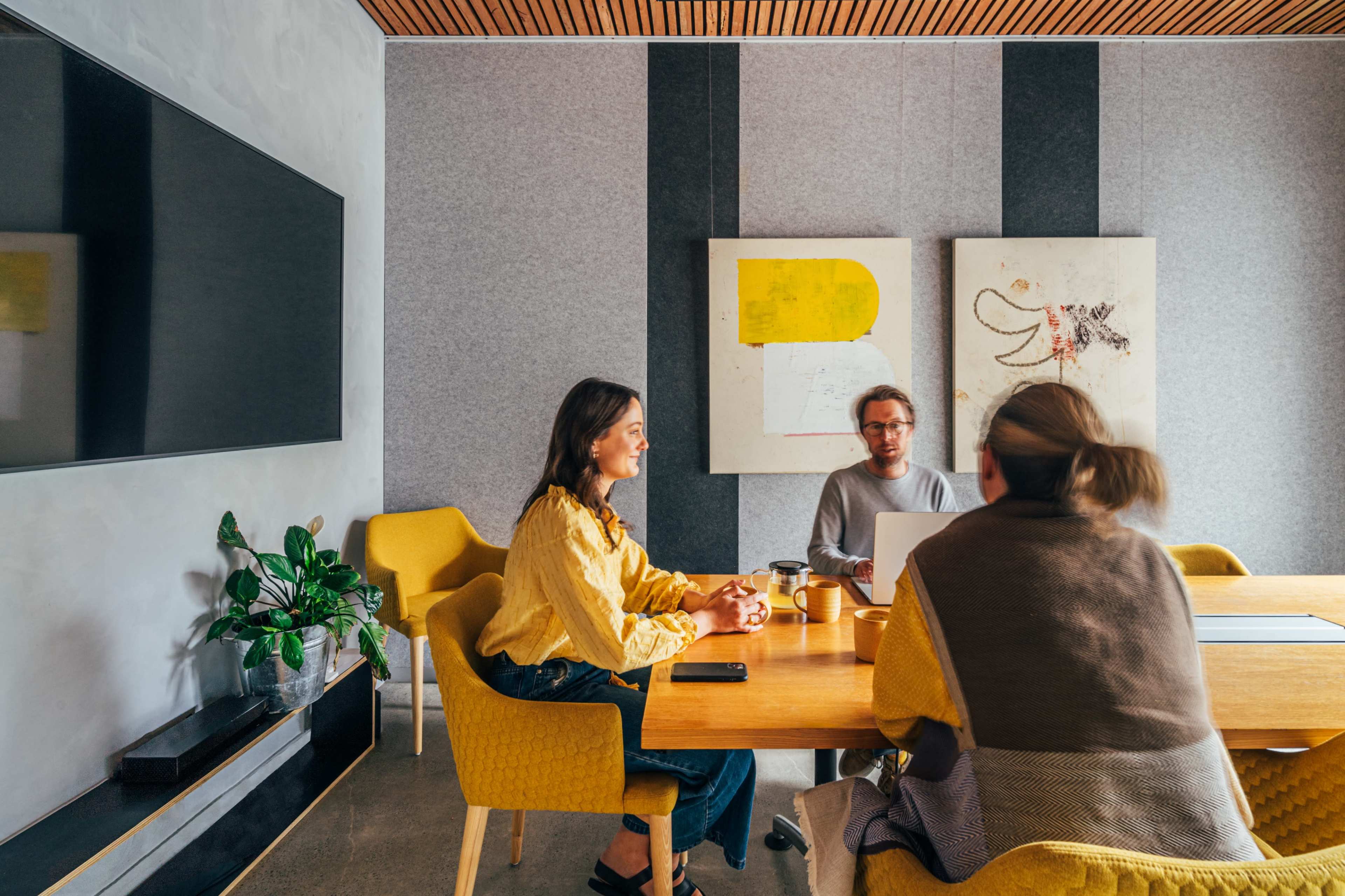 Three people engage in a discussion around a wooden table in a modern conference room, with yellow chairs and artwork on the walls.