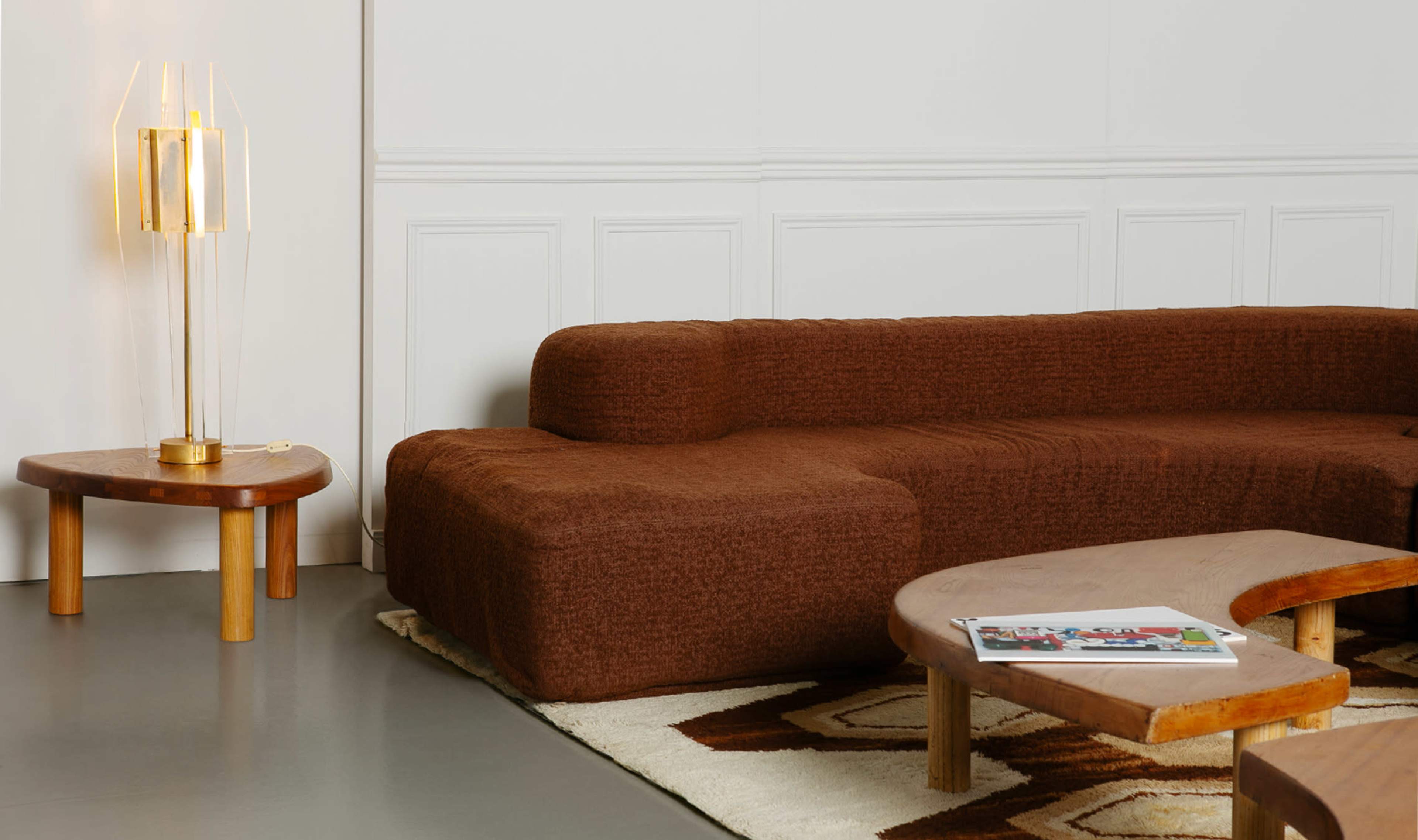 The image features a modern living room with a brown, curved sectional sofa, a round wooden side table with a lamp, and a low, irregularly shaped coffee table on a patterned rug.