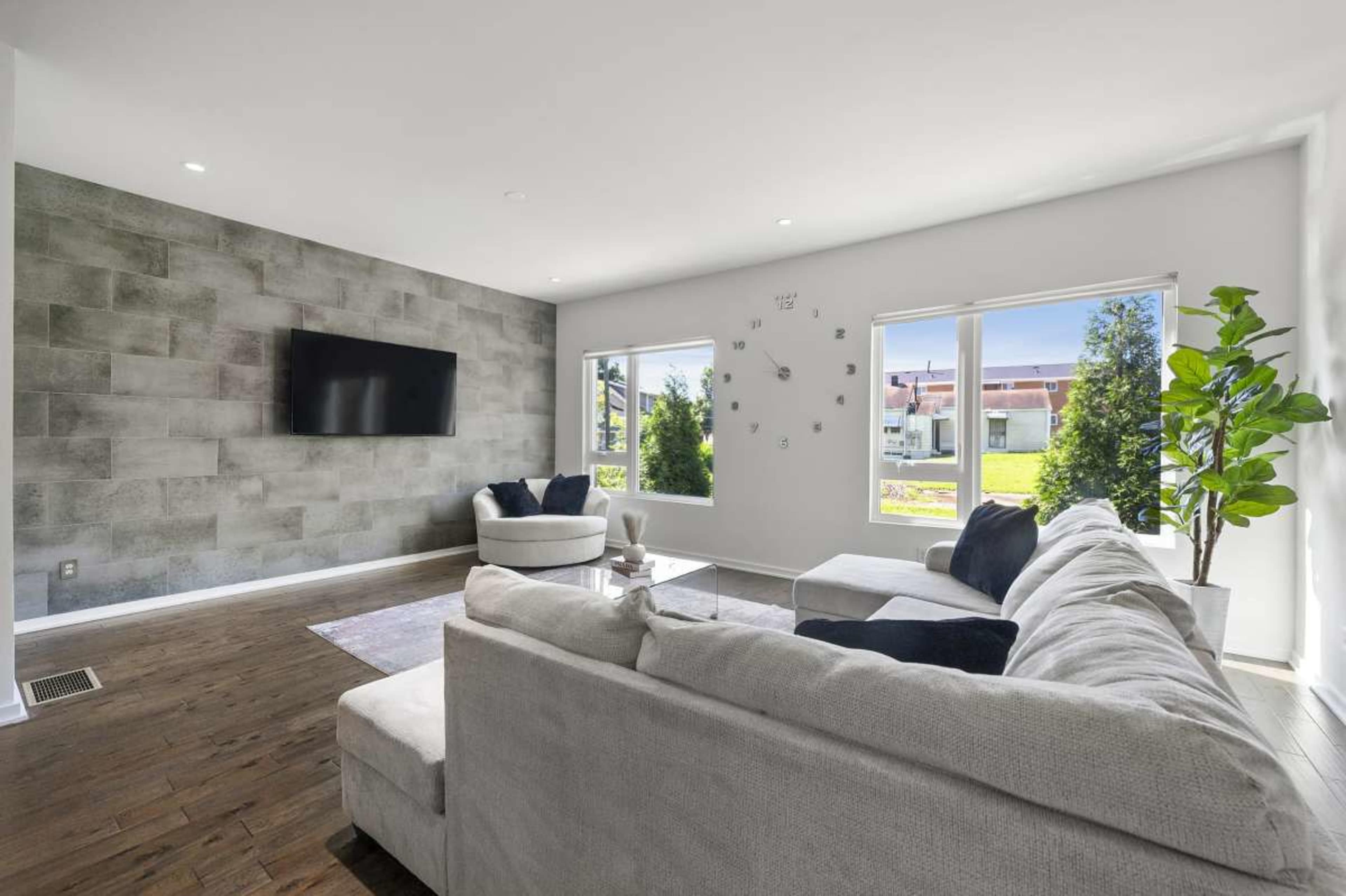 A bright living room features a light-colored sofa, a coffee table, and a large television mounted on a textured gray wall, with windows overlooking a garden.
