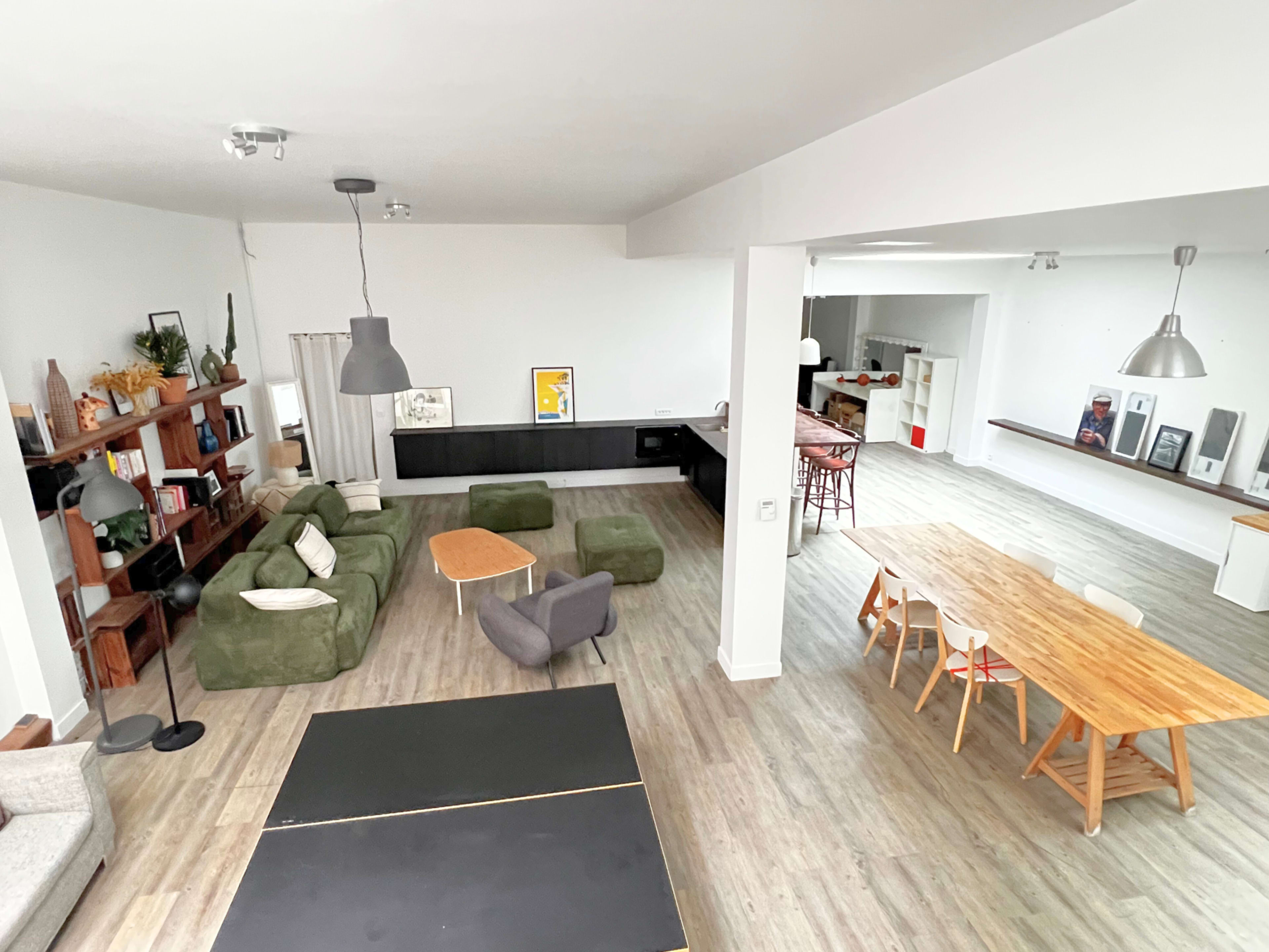 The image shows a spacious, modern living area featuring a green sectional sofa, a dining table, and various seating options in an open-plan design.