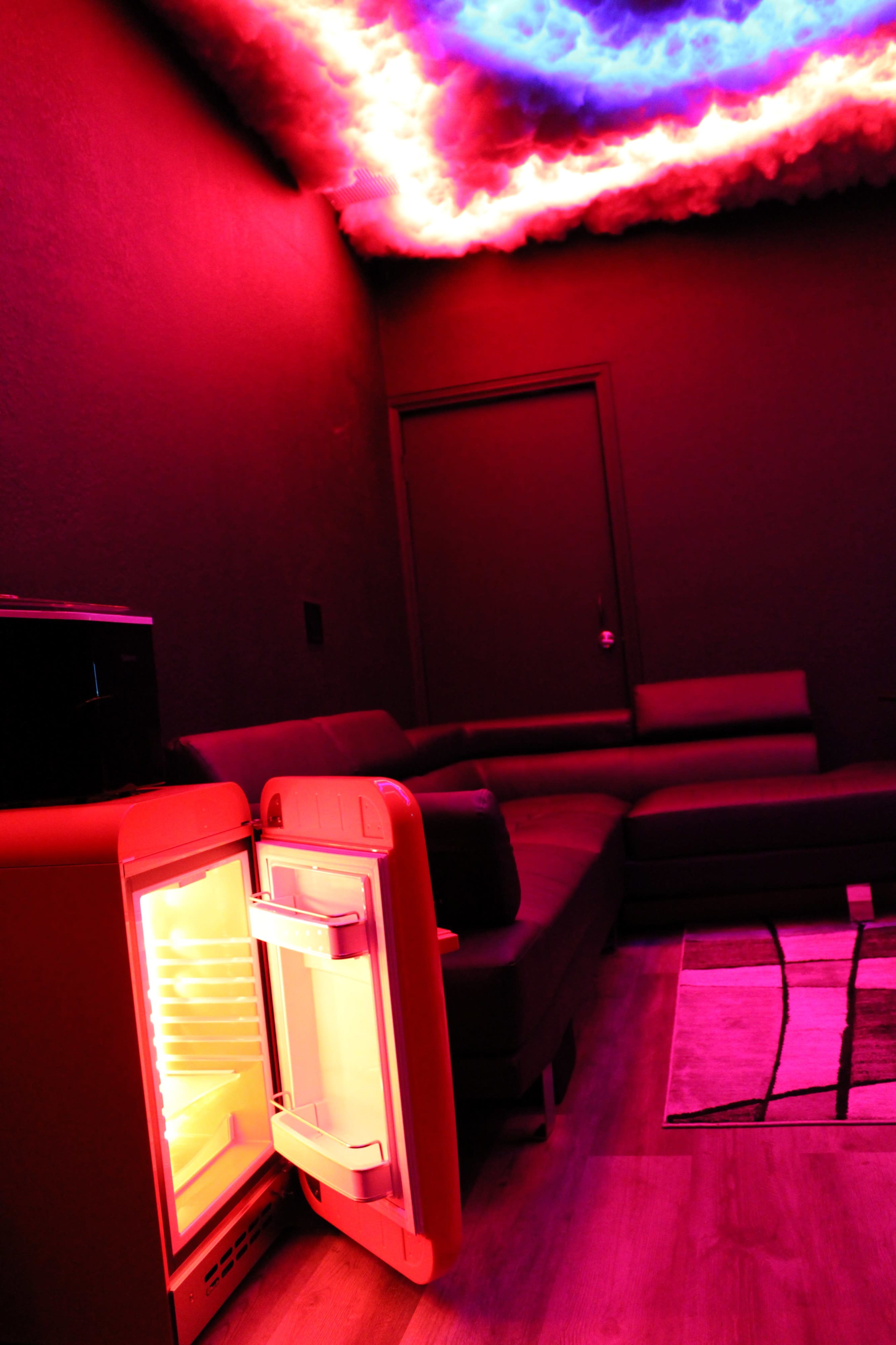 A brightly lit refrigerator with its door open is positioned in a dimly lit room featuring a sectional couch and a ceiling illuminated with a colorful cloud-like design.