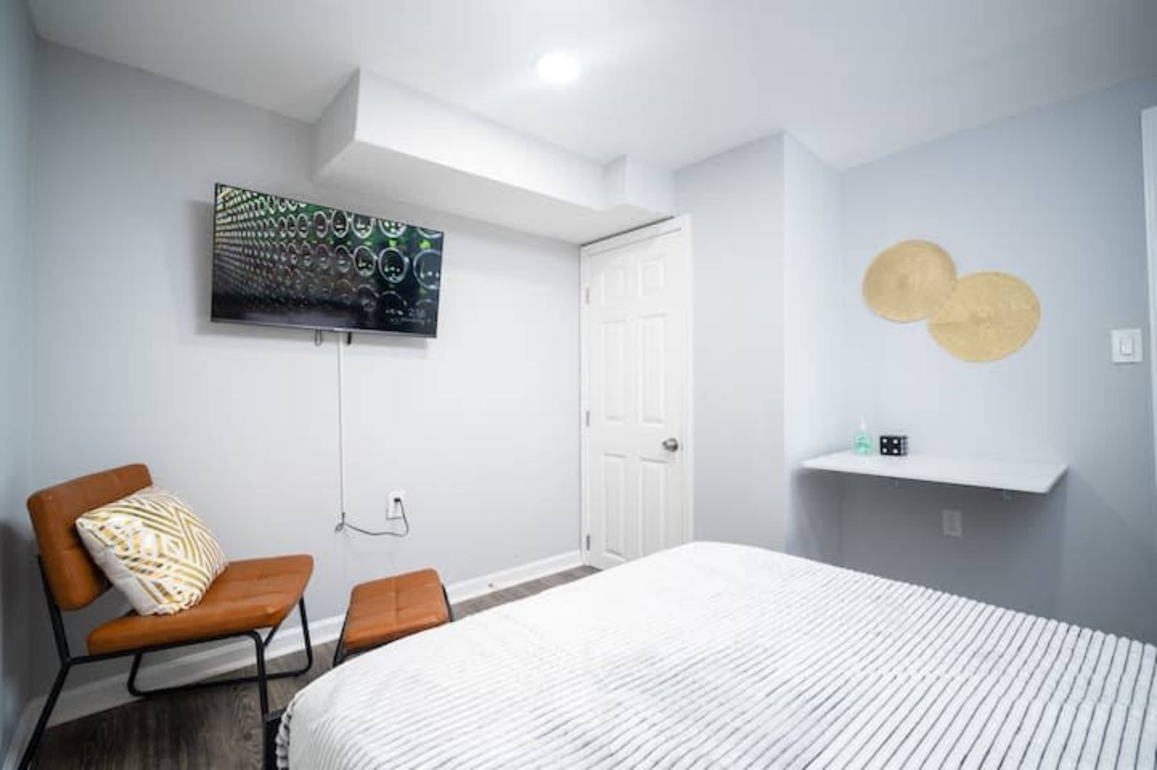 A small bedroom with a bed, a television mounted on the wall, a brown chair, and a white side table.