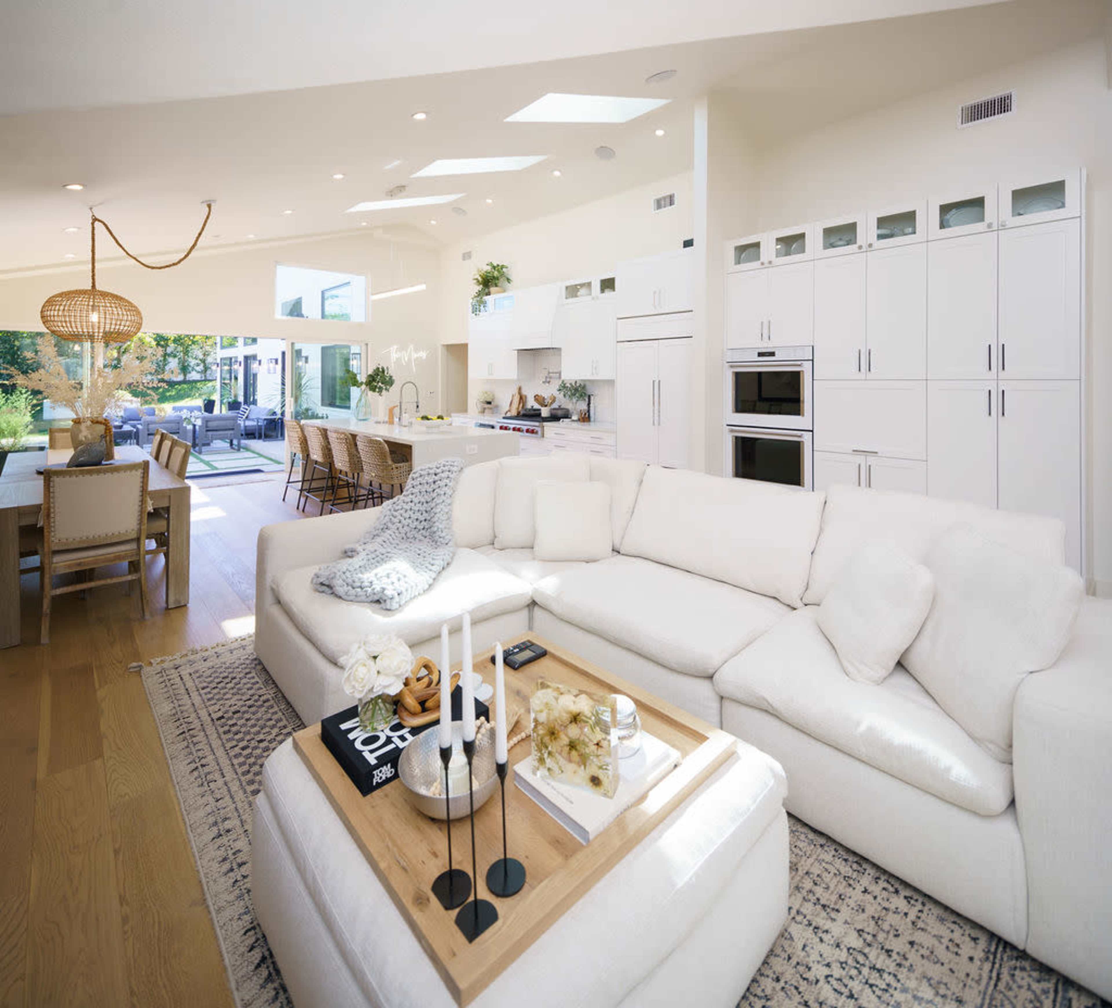 A modern, bright living room with a large white sectional sofa, a wooden coffee table adorned with decorative items, and an open kitchen area in the background.