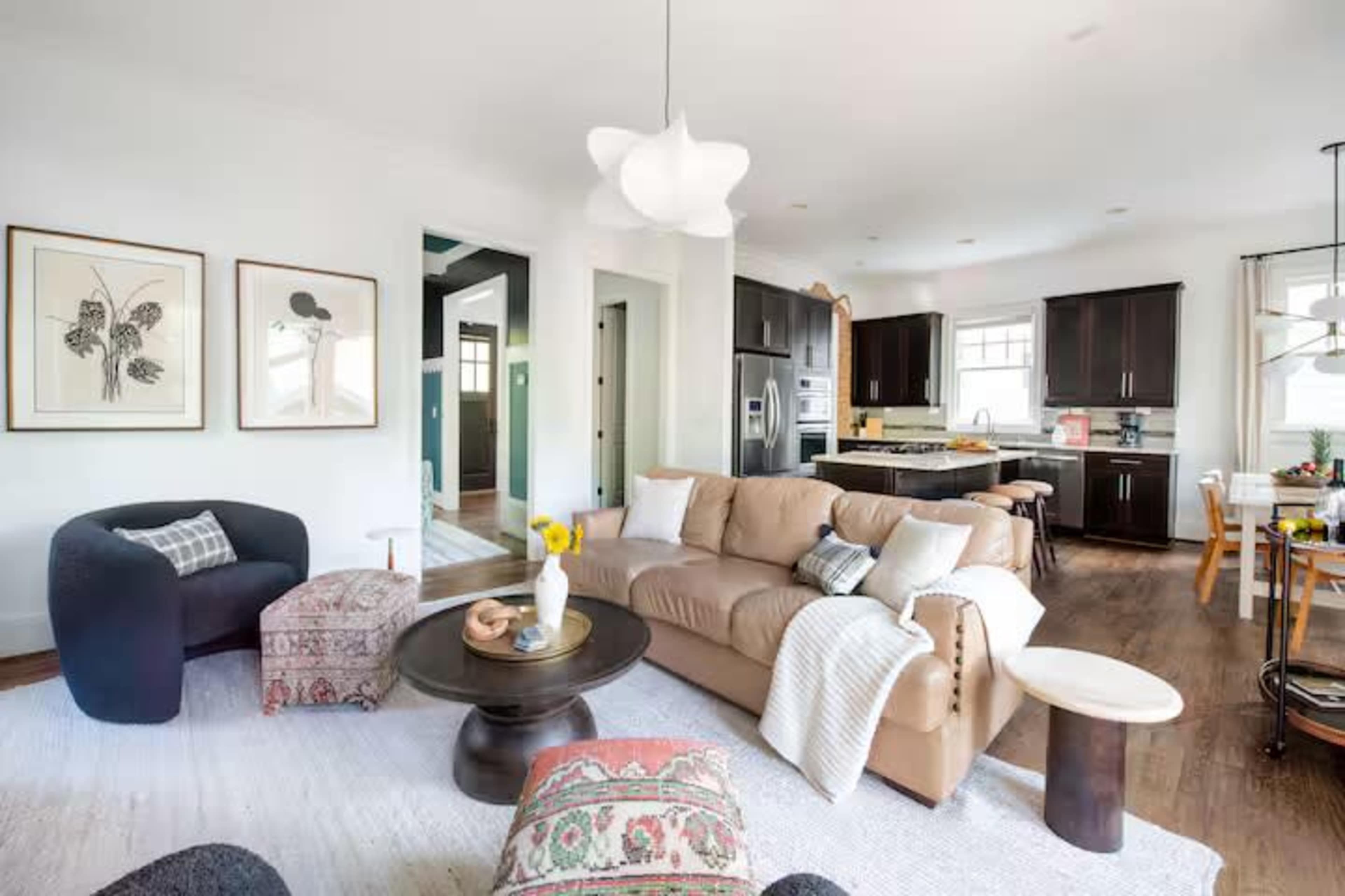 The image shows a modern living room with a light-colored sofa, various seating options, and an open kitchen in the background.