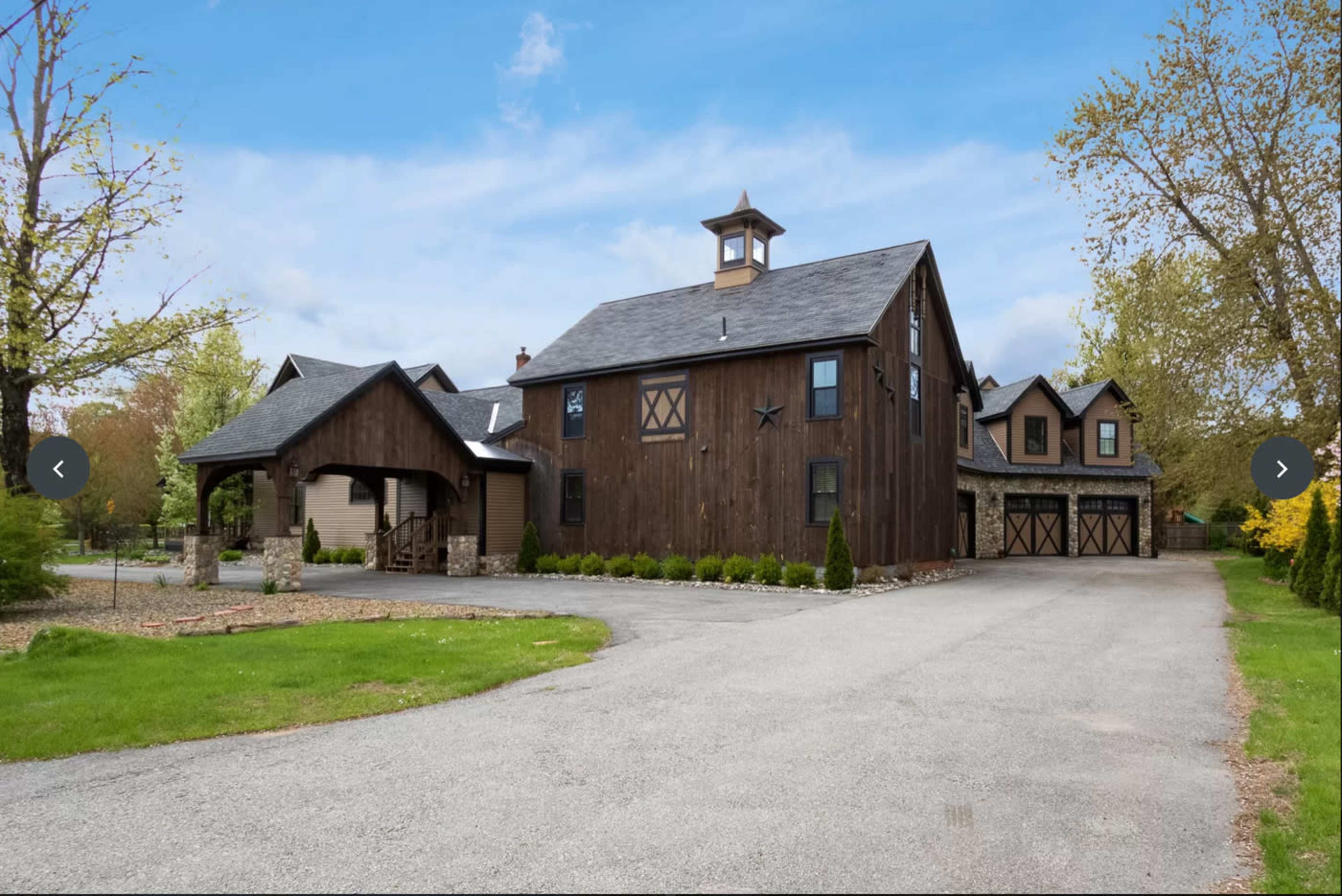 A large wooden barn-style house with a cupola, surrounded by a gravel driveway and landscaped greenery.