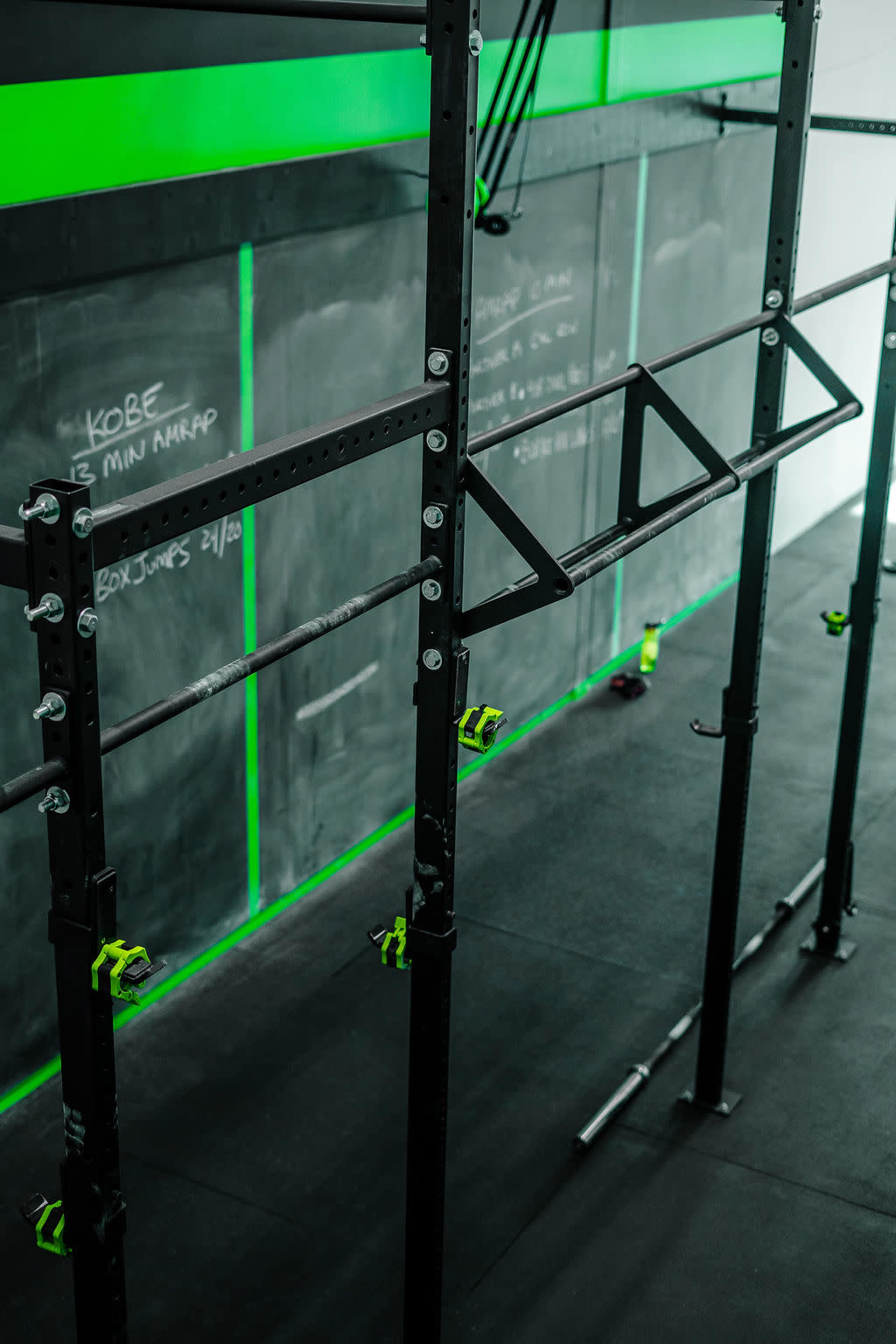 A gym space features a metal rig with green accents and a chalkboard wall displaying workout information.