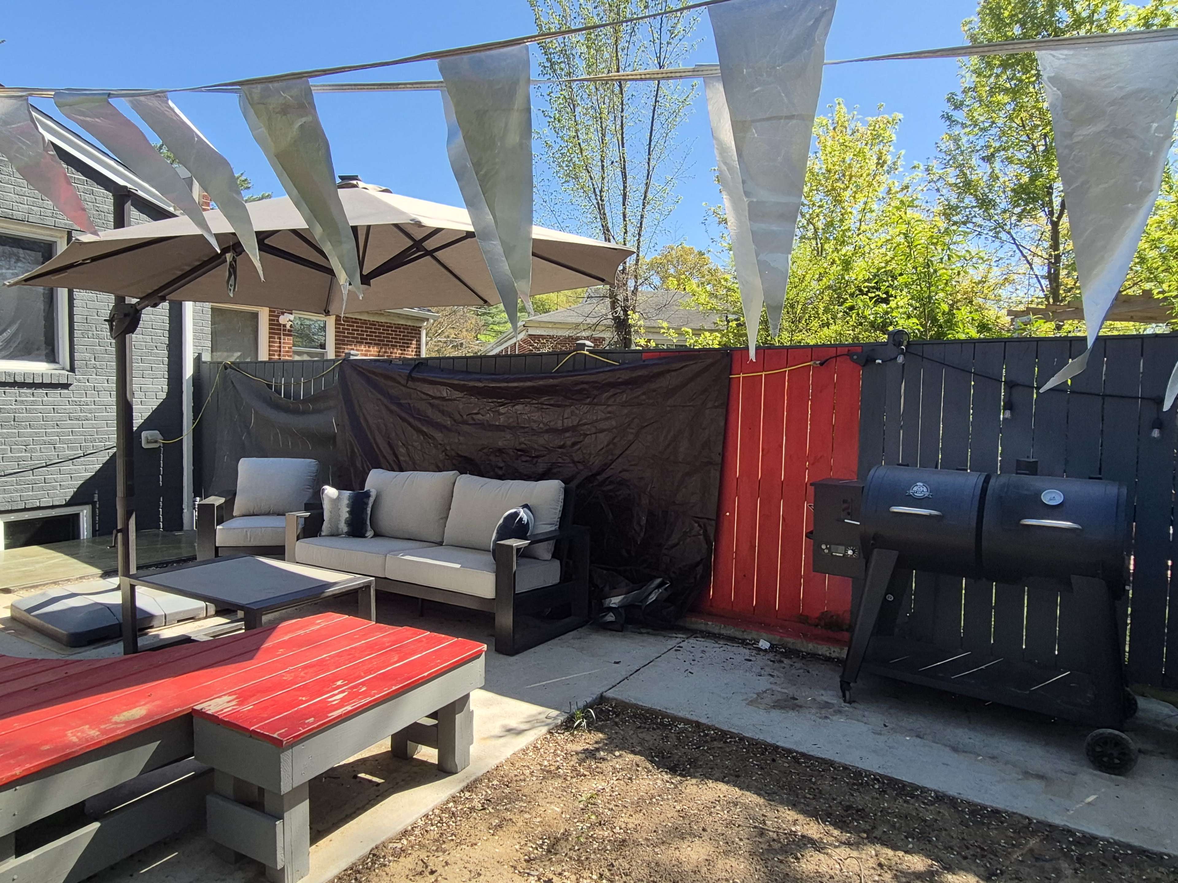 A patio area features a red and black fence, a gray umbrella over a seating arrangement, and a grill beside a red picnic table.