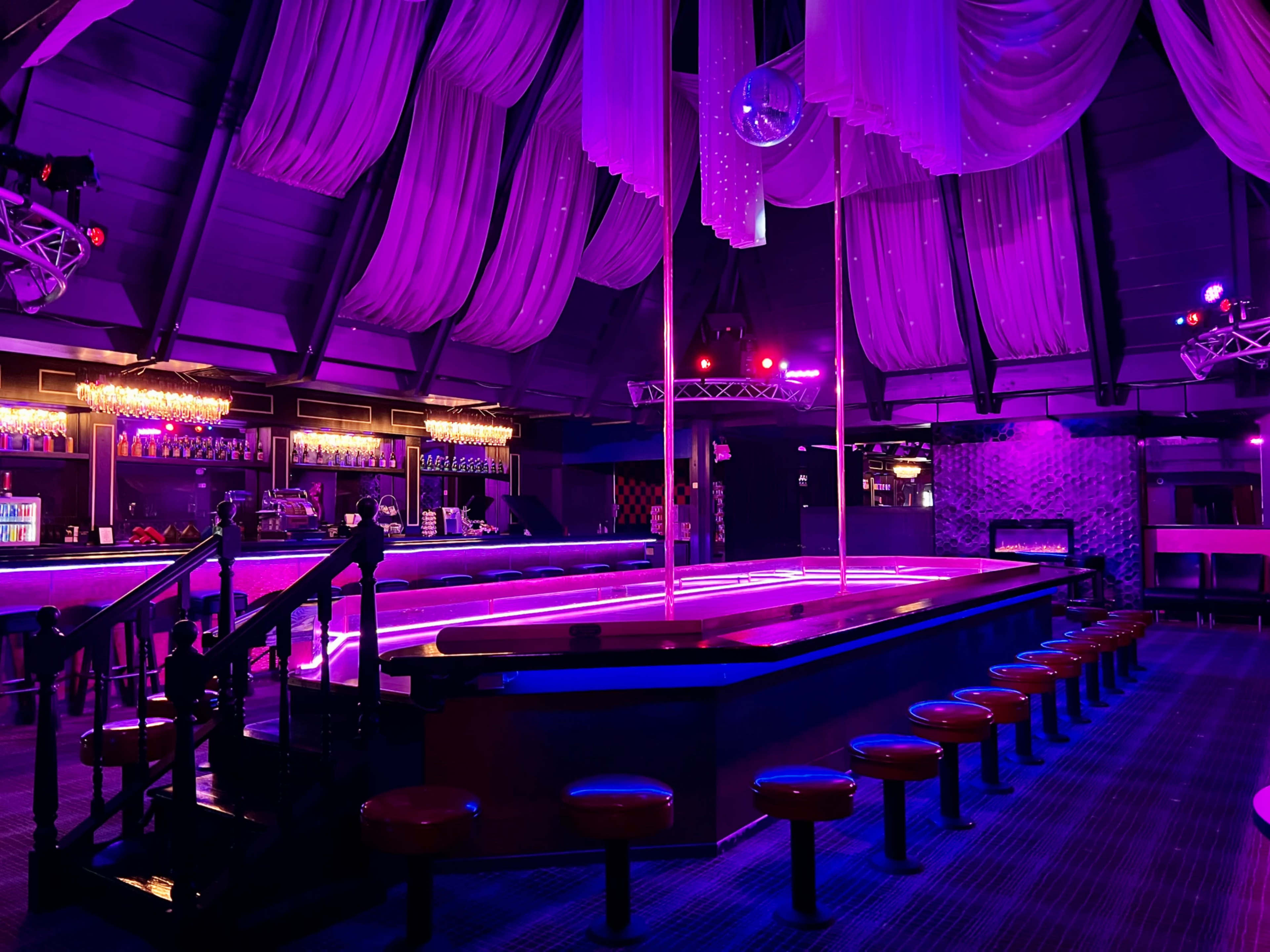 A dimly lit nightclub interior with purple lighting, featuring a bar area and a central stage surrounded by seating.