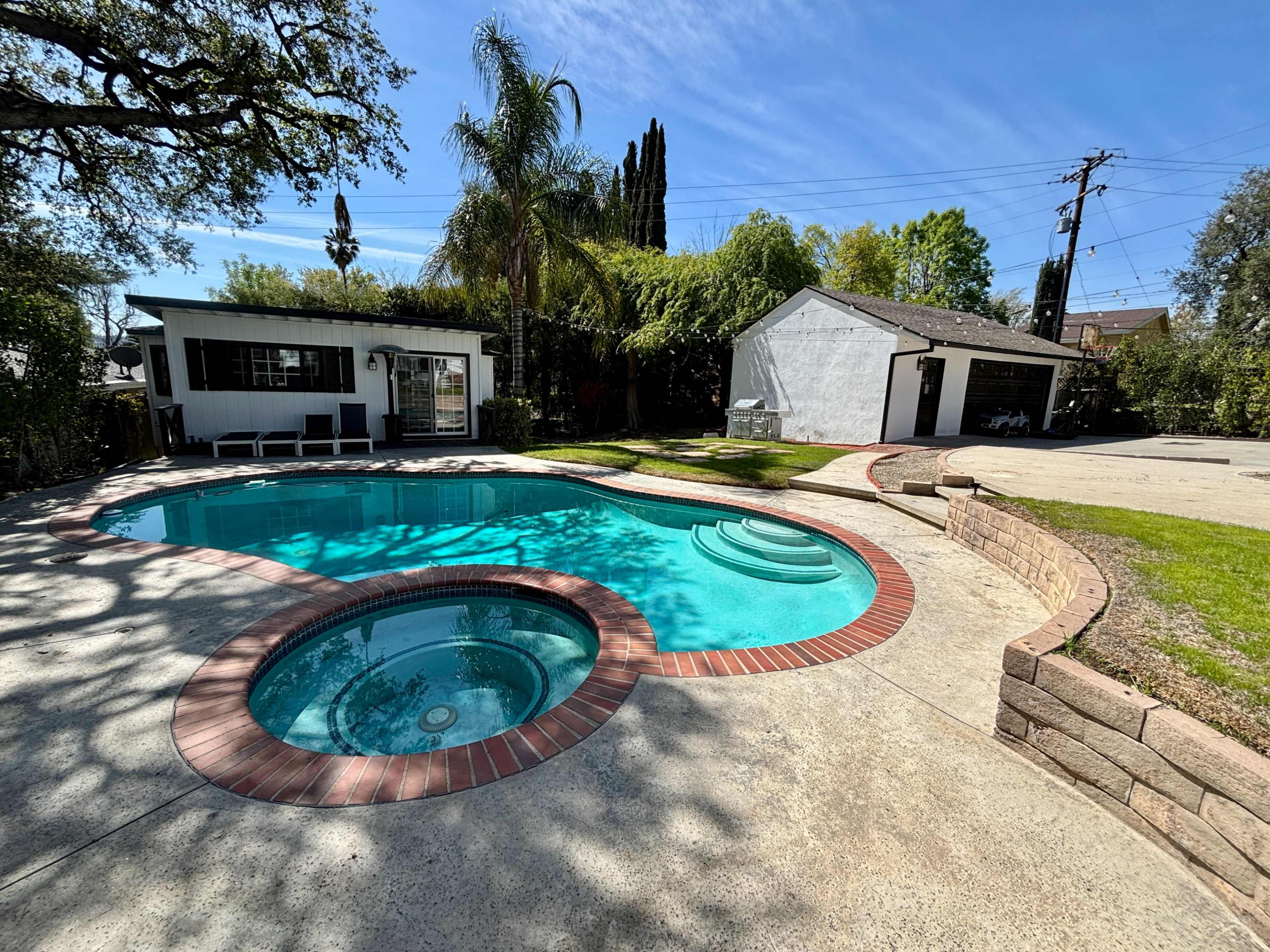 The image shows a clear blue swimming pool with a spa, surrounded by concrete and grass, adjacent to a white building and tropical trees.