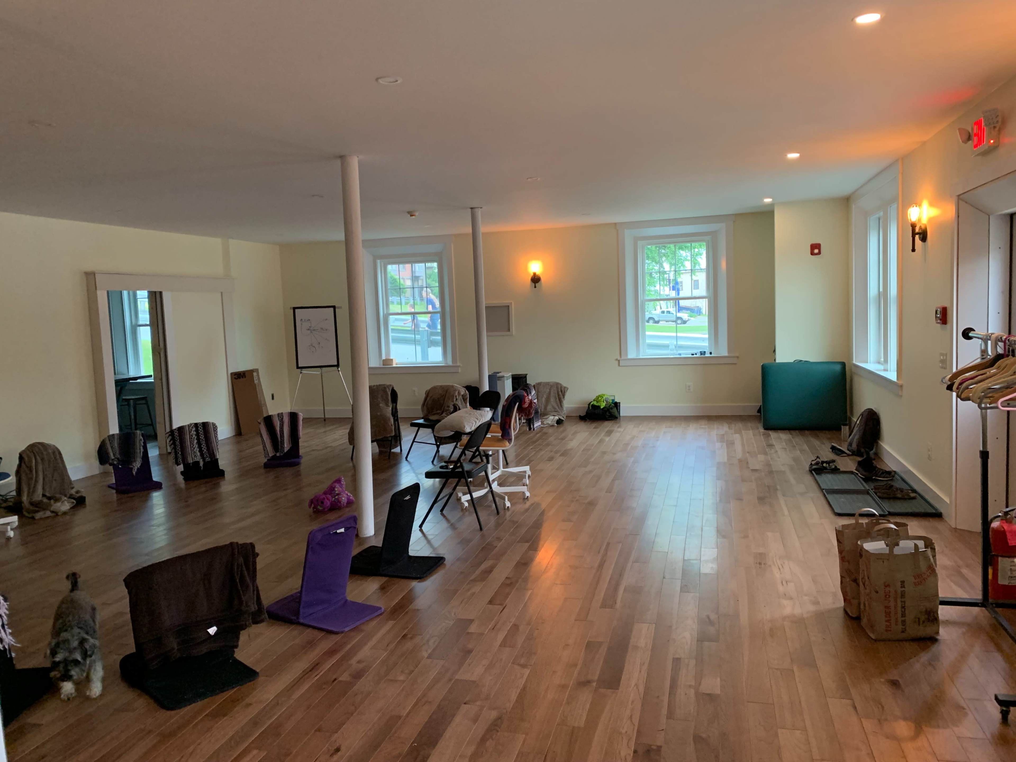 The image shows a spacious room with wooden flooring, scattered chairs, and various items placed around the area, including bags and mats.