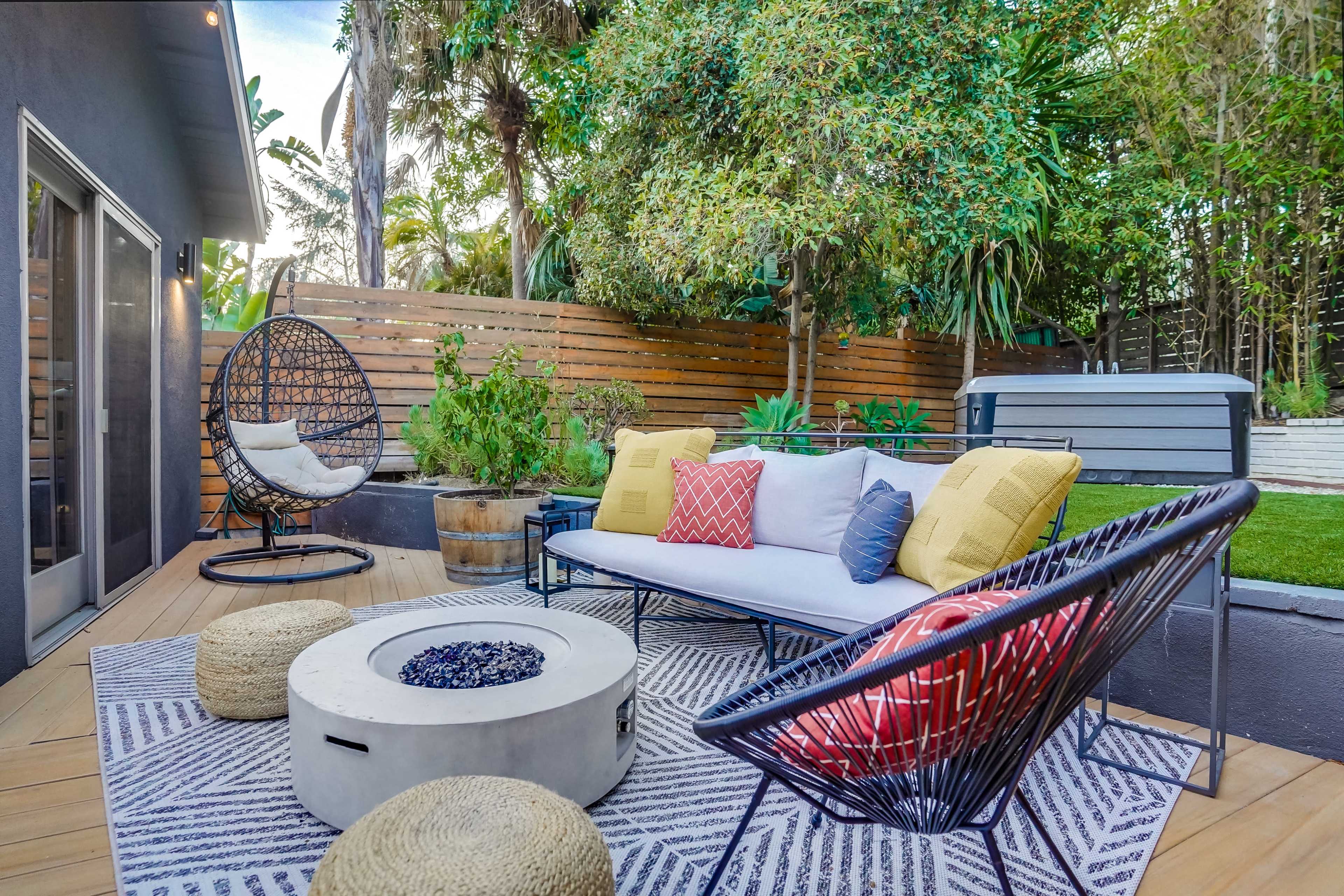 A stylish backyard patio featuring a sofa set, a fire pit, woven chairs, and lush greenery.