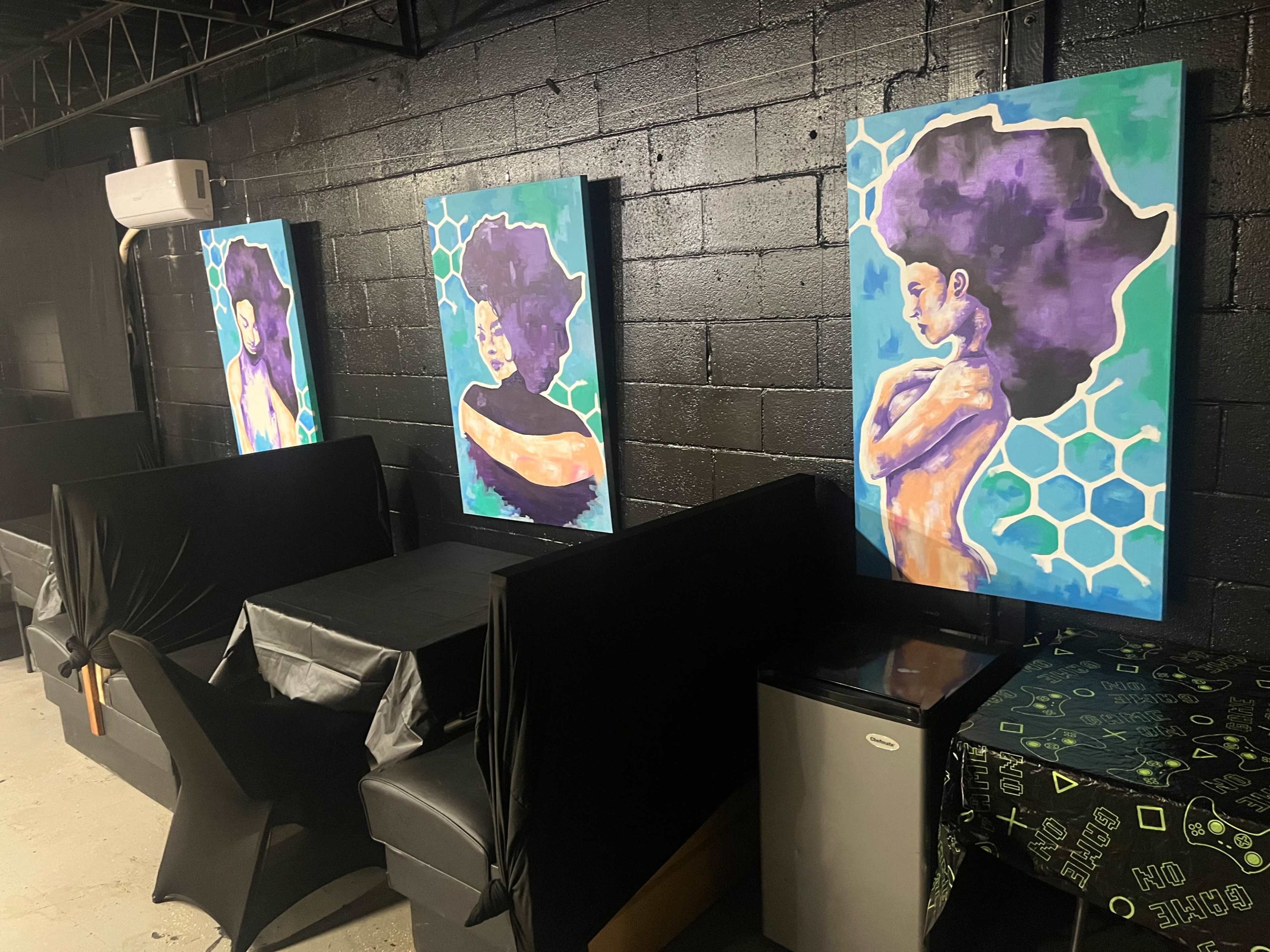 The image shows a black-walled room with four large, colorful paintings of women on the walls and various seating arrangements, including black booths and a table covered in a green tablecloth.
