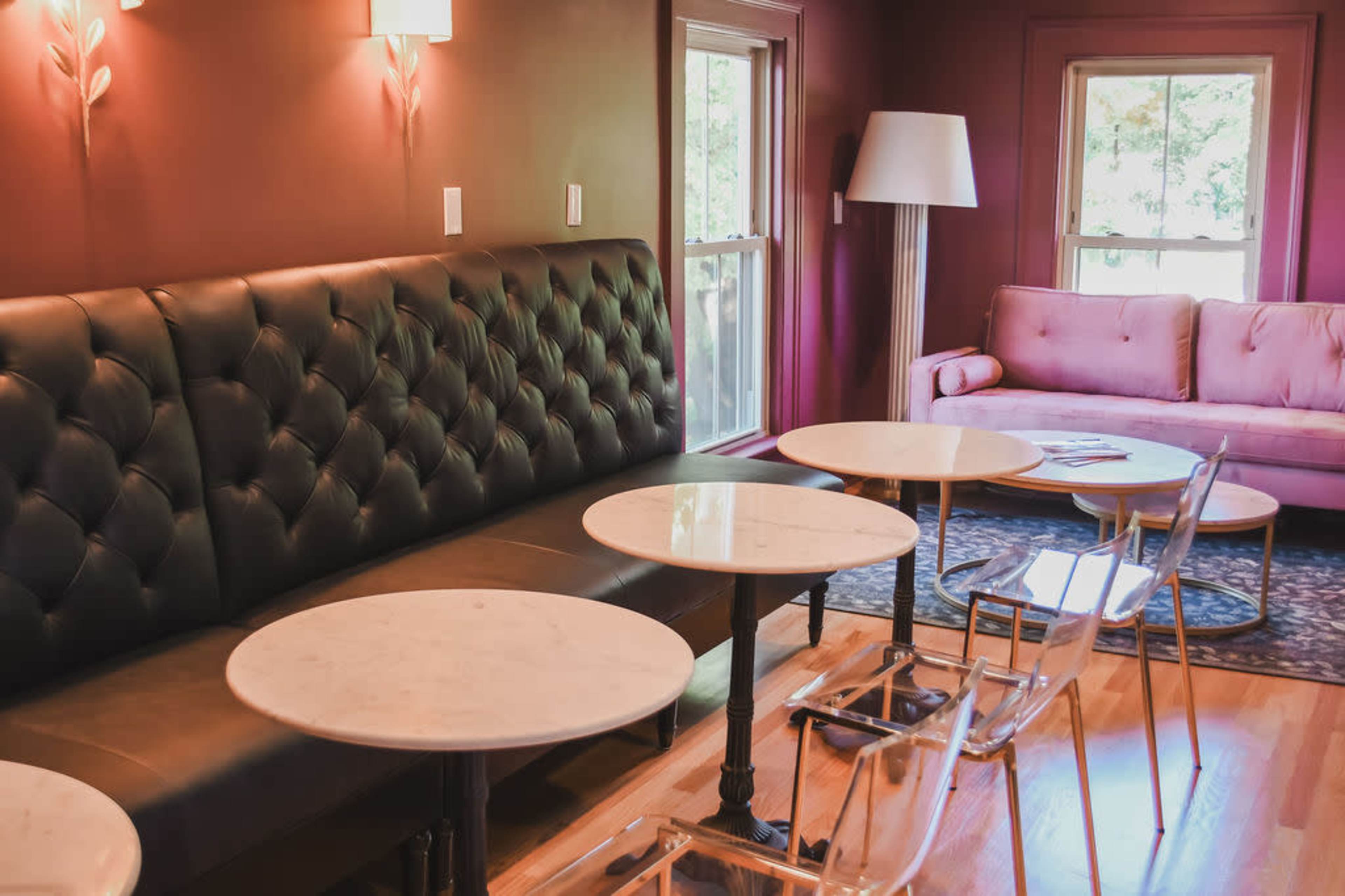 The image shows a cozy coffee shop interior featuring a black tufted bench, light-colored marble-top tables, a pink sofa, and large windows allowing natural light.