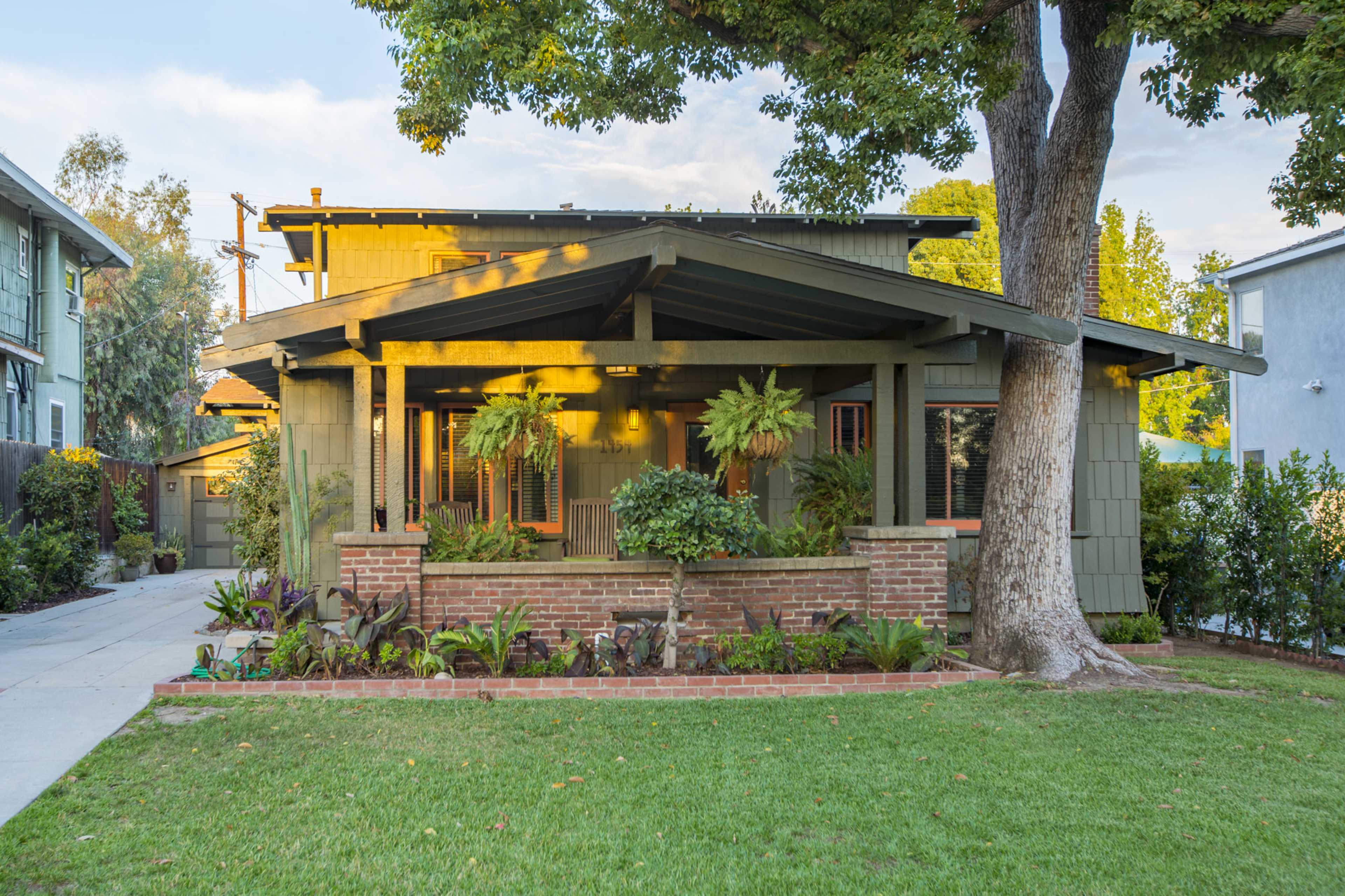 A single-story house with a sloped roof, surrounded by greenery and a brick planter, features a welcoming porch with seating.