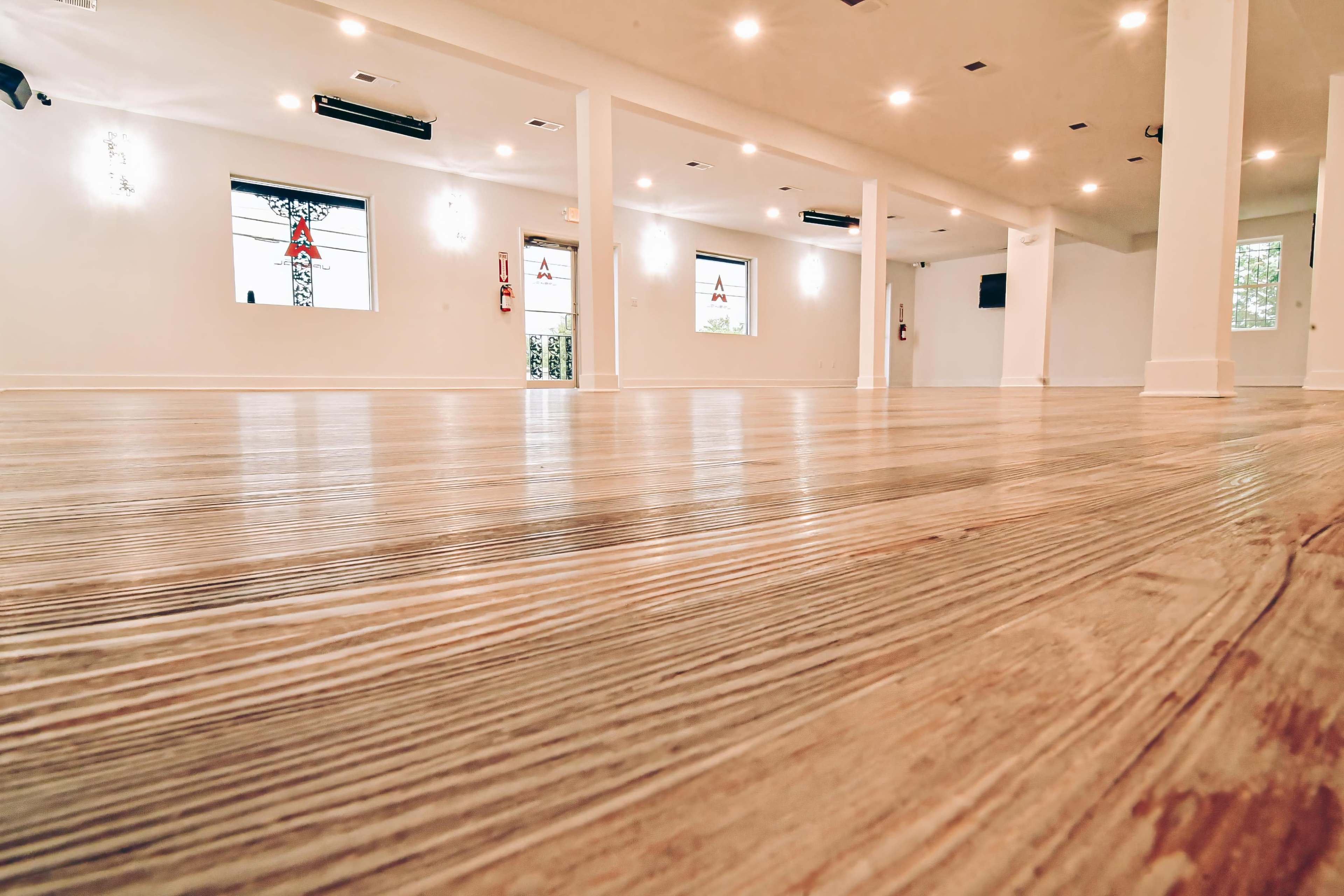 The image shows an empty room with wooden flooring and white walls, illuminated by overhead lights.