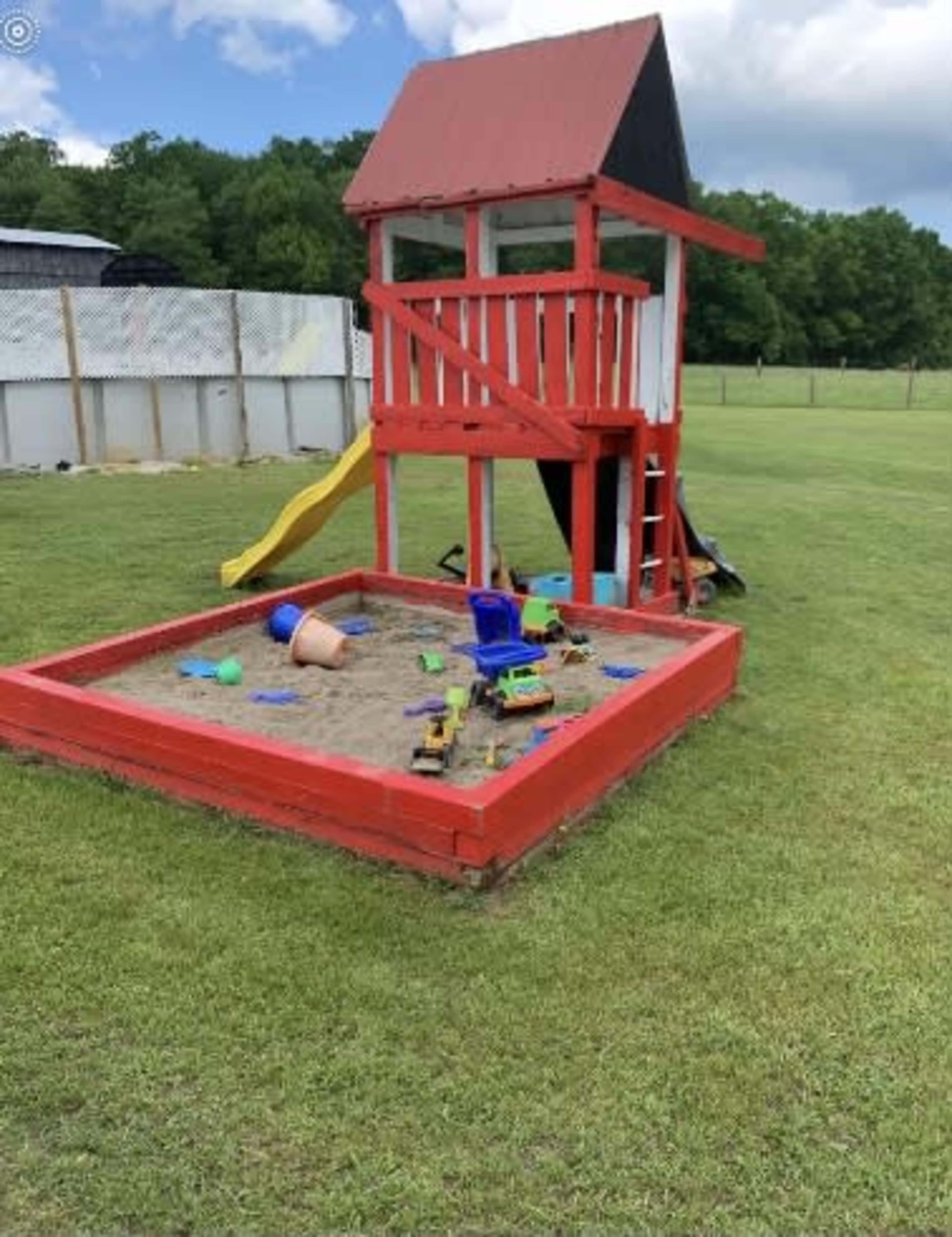 The image shows a red wooden playhouse with a slide beside a sandpit filled with toys, set in a grassy field.