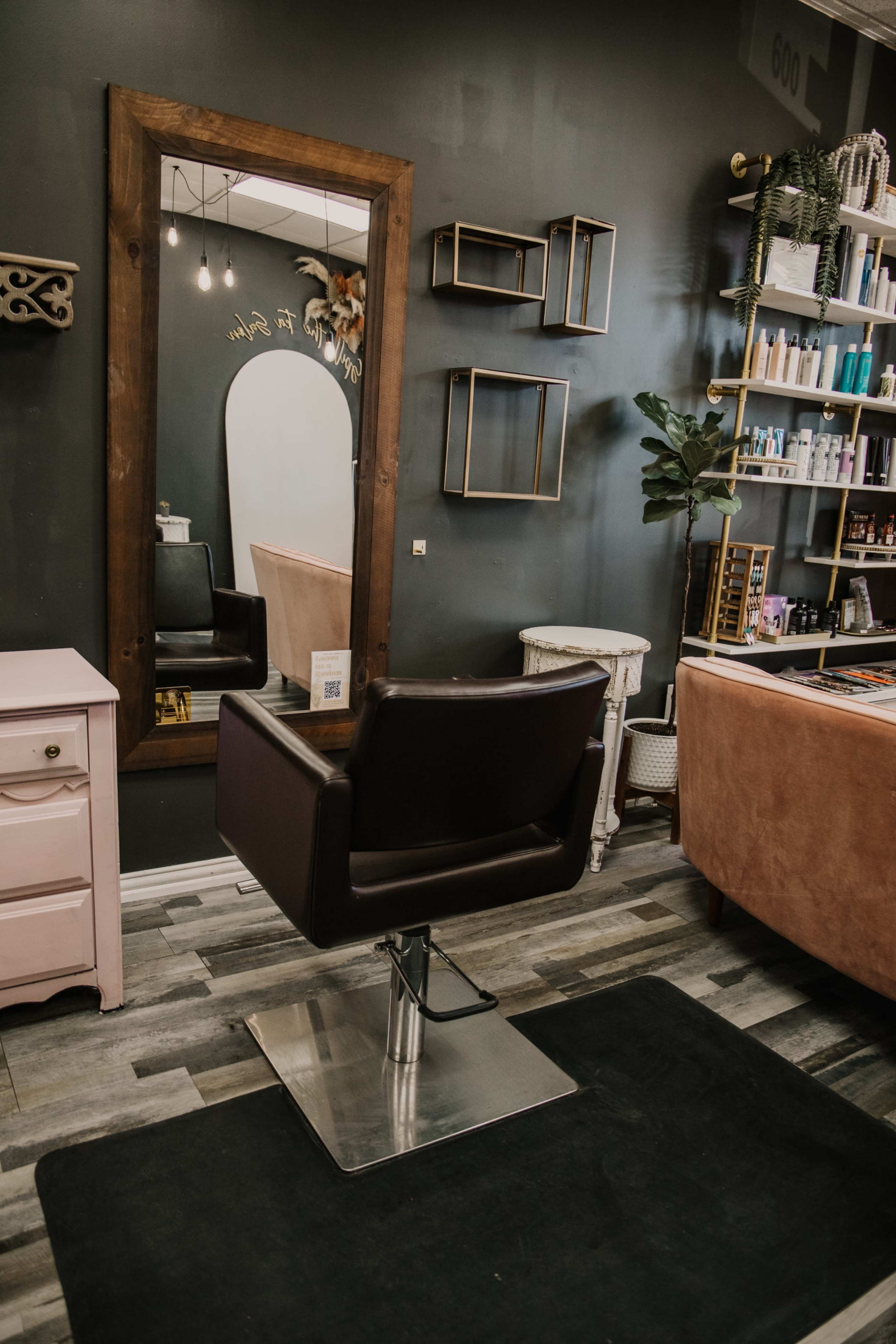 A hair salon interior featuring a black styling chair in front of a large mirror, with shelves of hair products and a decorative plant against a dark wall.