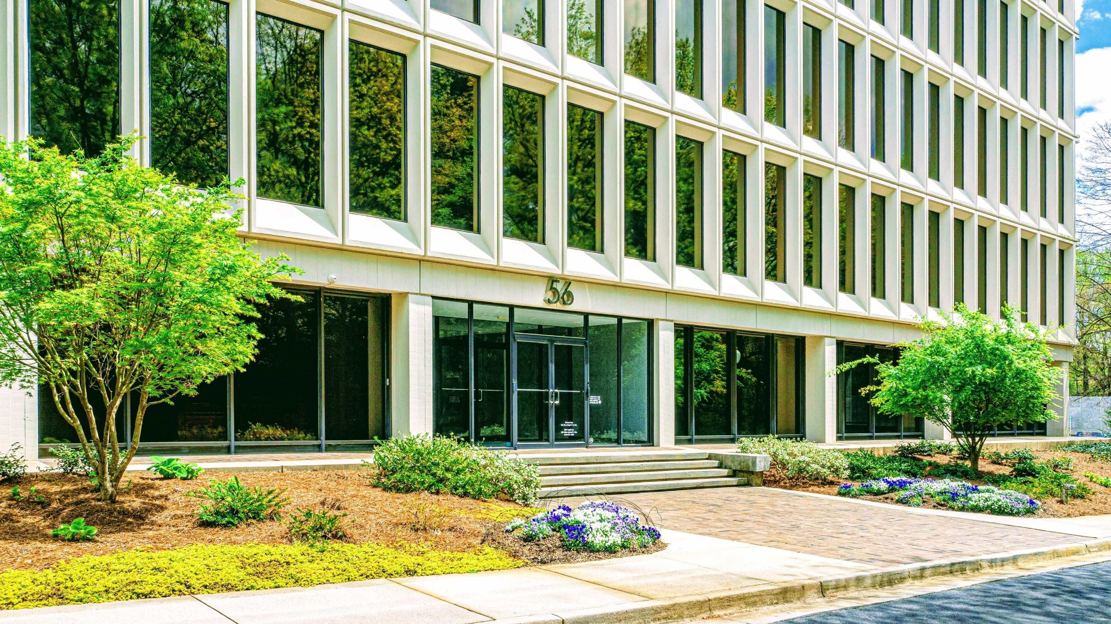 The entrance of a modern office building with large glass windows and a landscaped pathway lined with flowers and greenery.