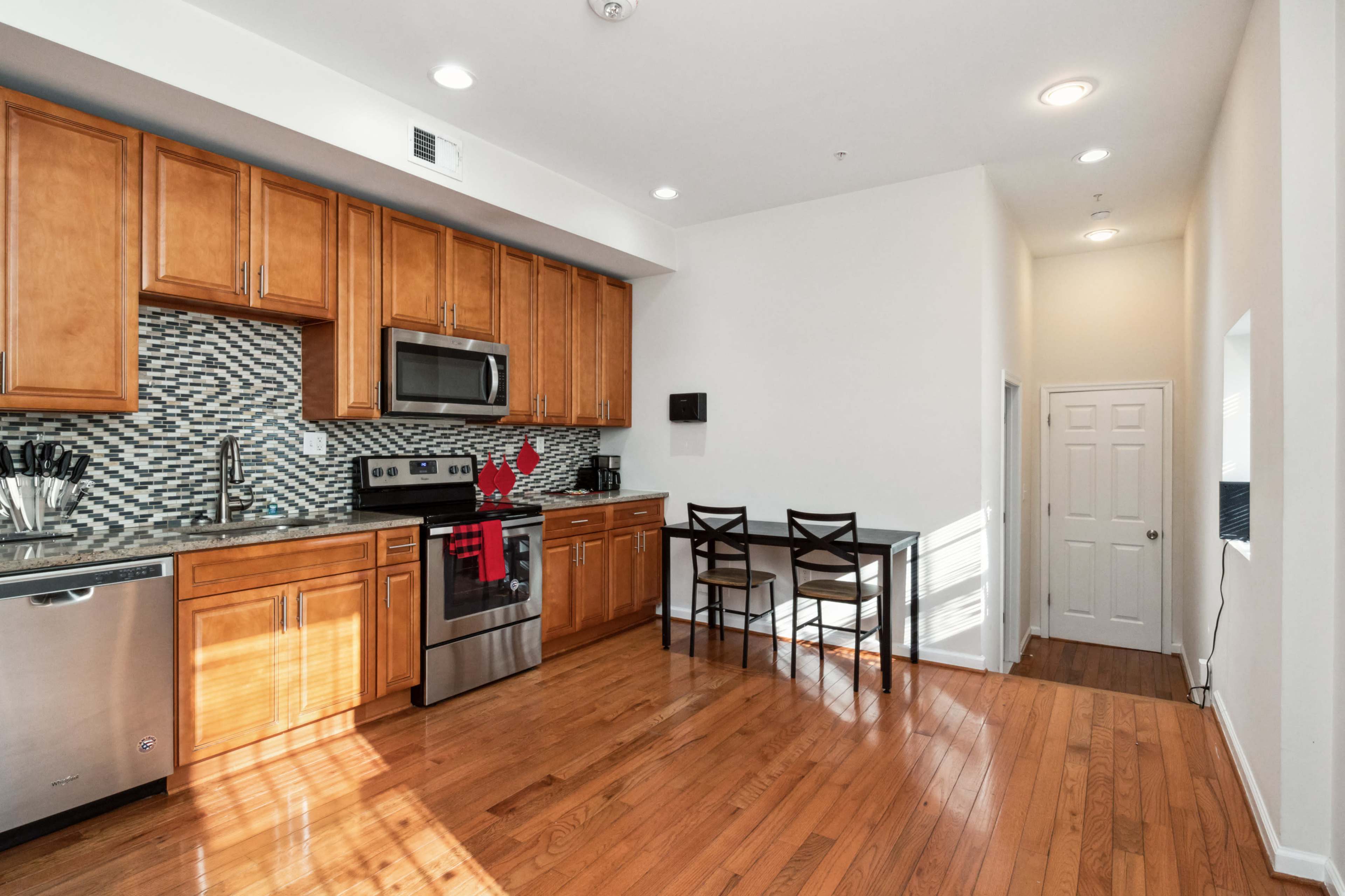 A modern kitchen features wooden cabinets, a stainless steel stove and dishwasher, and a small dining table with chairs against a brightly lit wall.
