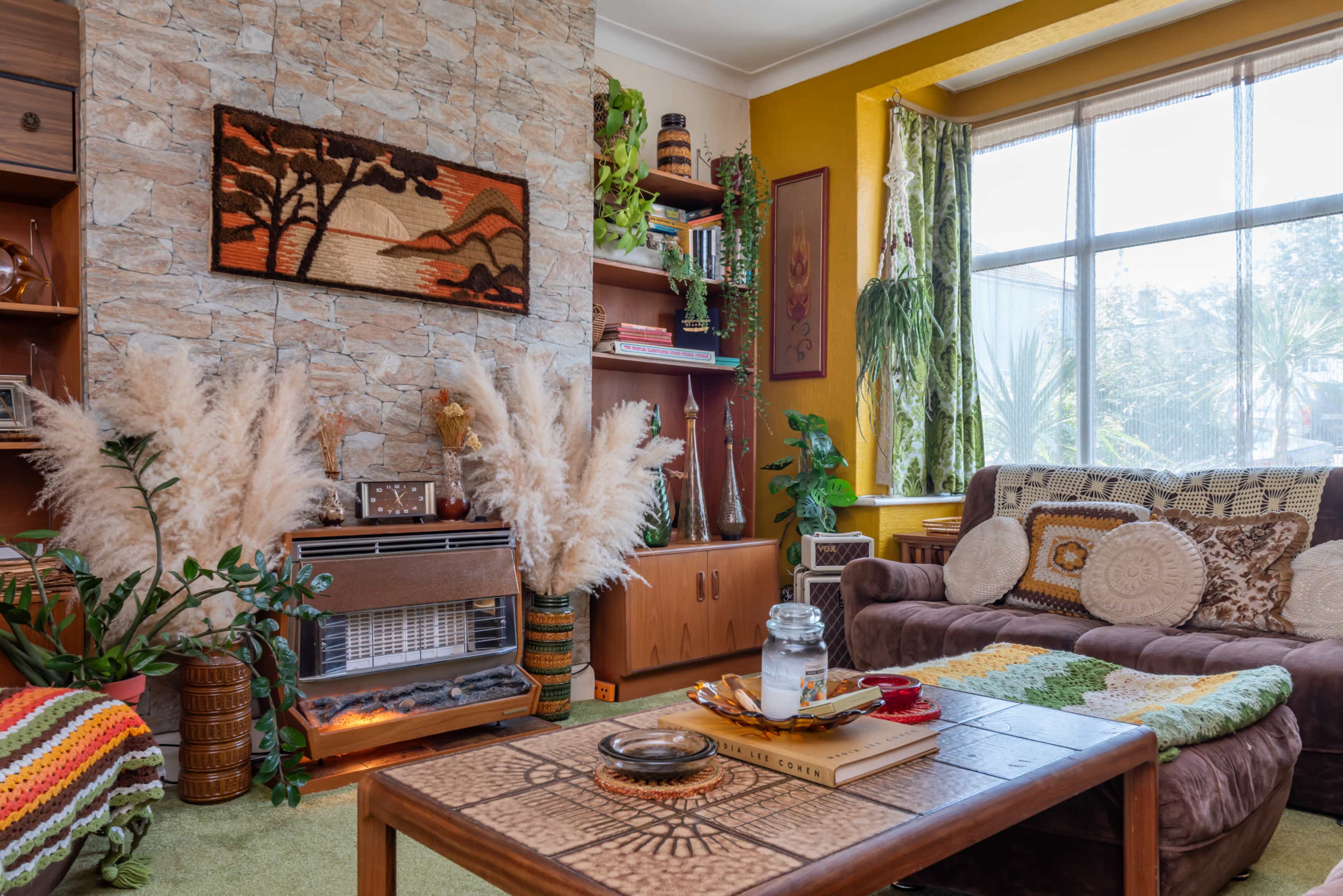 The image depicts a cozy living room featuring a stone accent wall, a brown sofa, decorative plants, and a coffee table adorned with books and a jar.