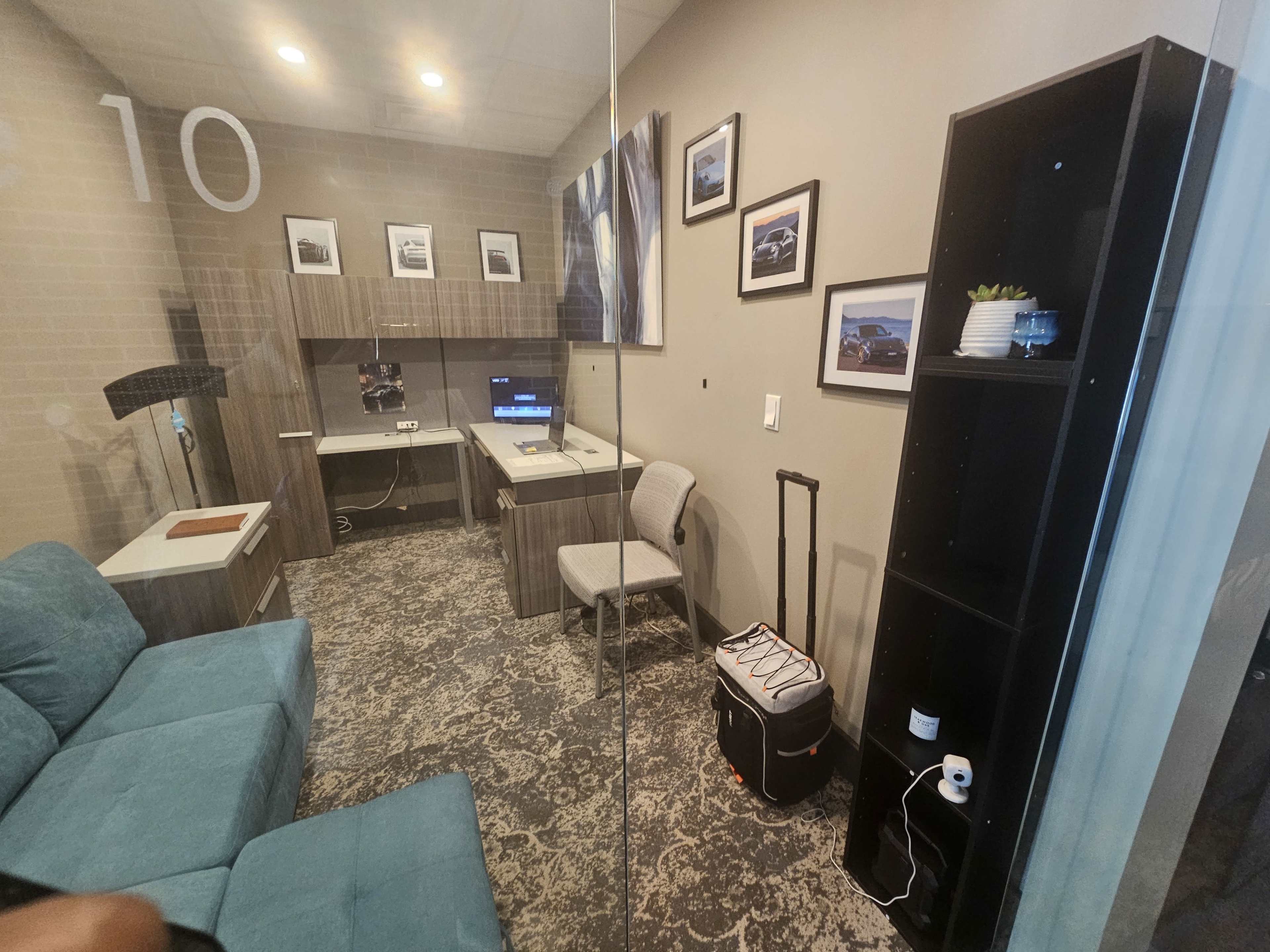 The image shows a small office room with a blue couch, a desk with a computer, a chair, a bookshelf, and a suitcase.