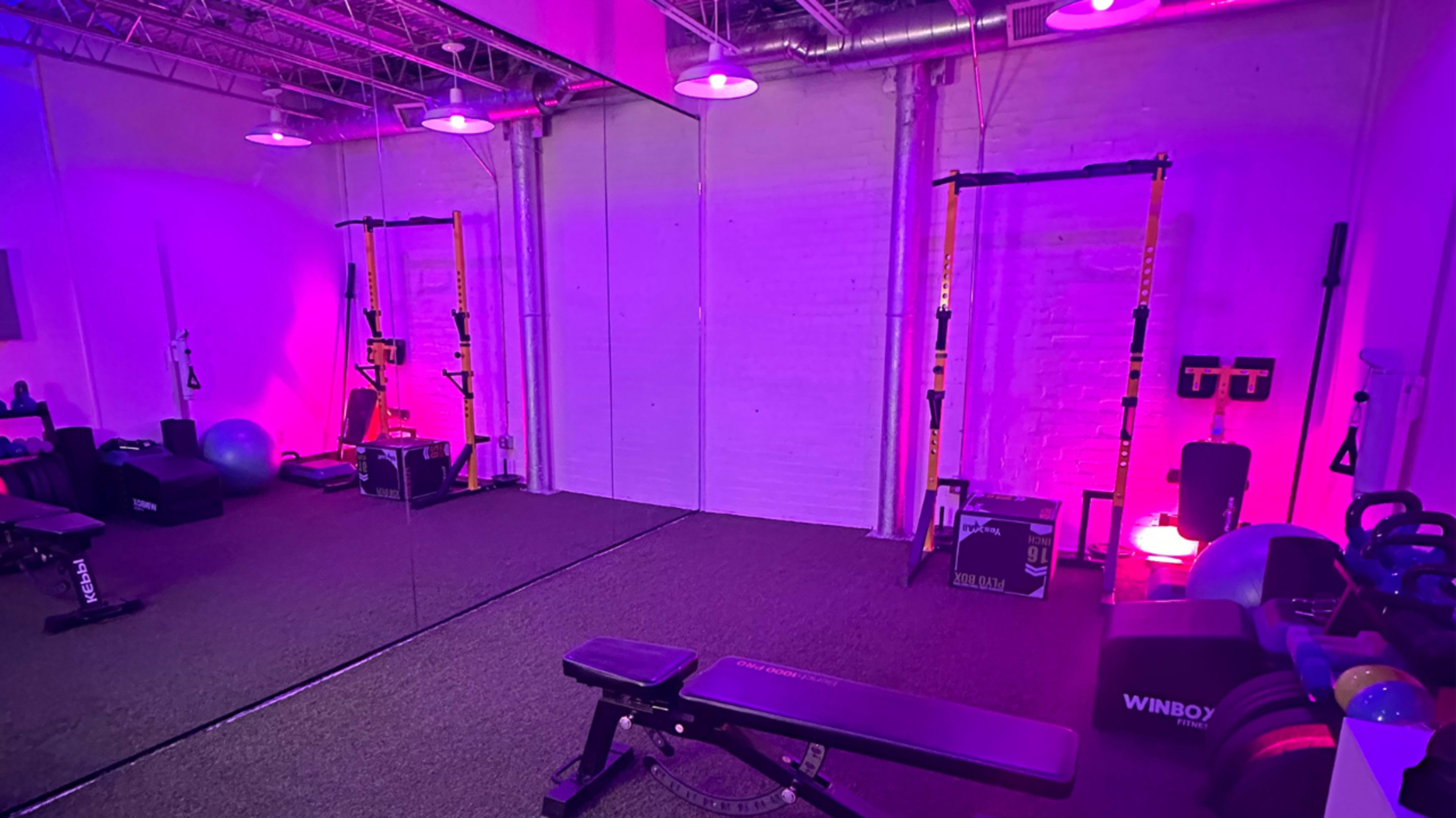 The image shows a gym space with exercise equipment, including a bench and weights, surrounded by purple lighting and a large mirror.