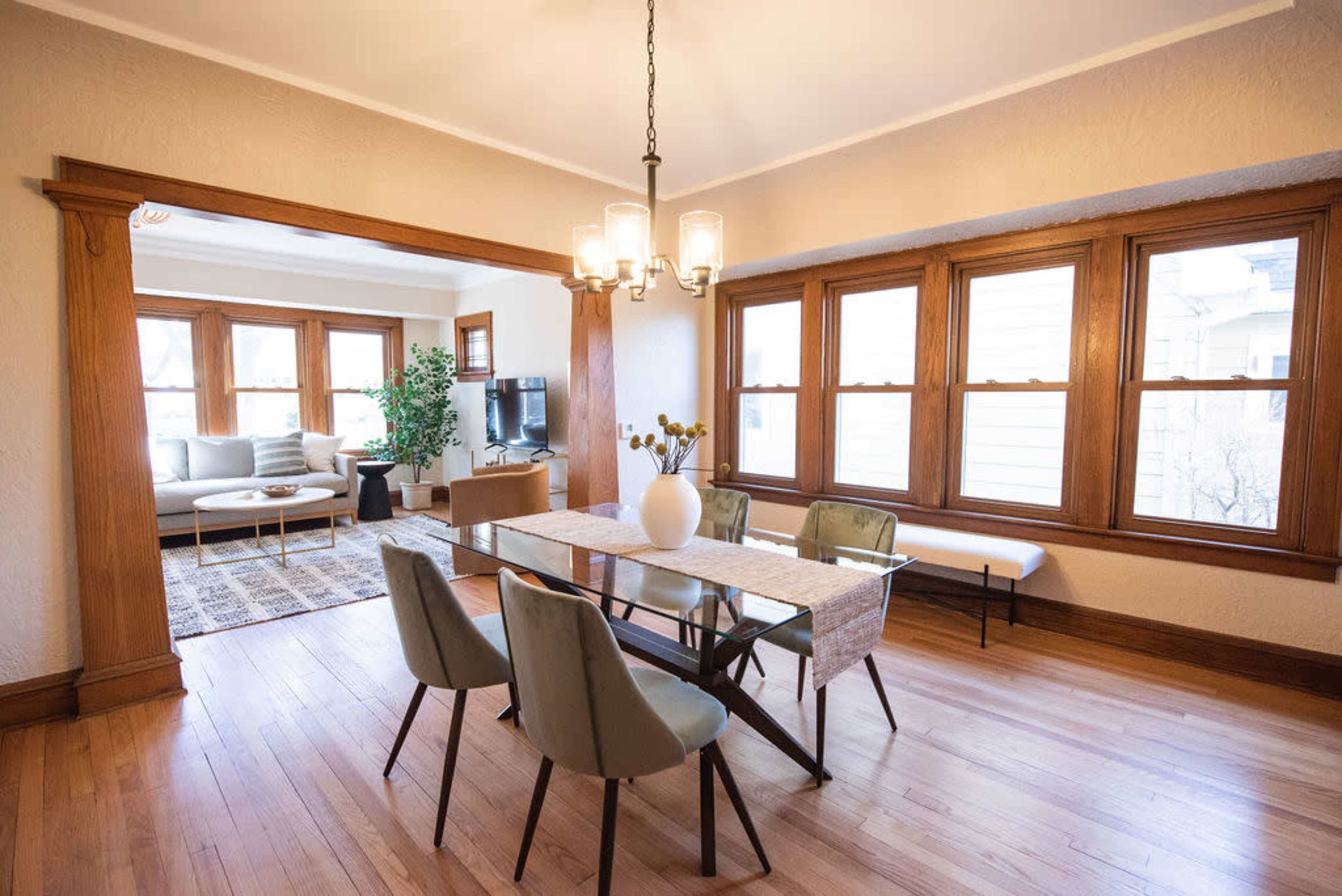 A dining area features a rectangular table with four chairs, a vase on the table, and a view into a connected living space with large windows and a sofa.