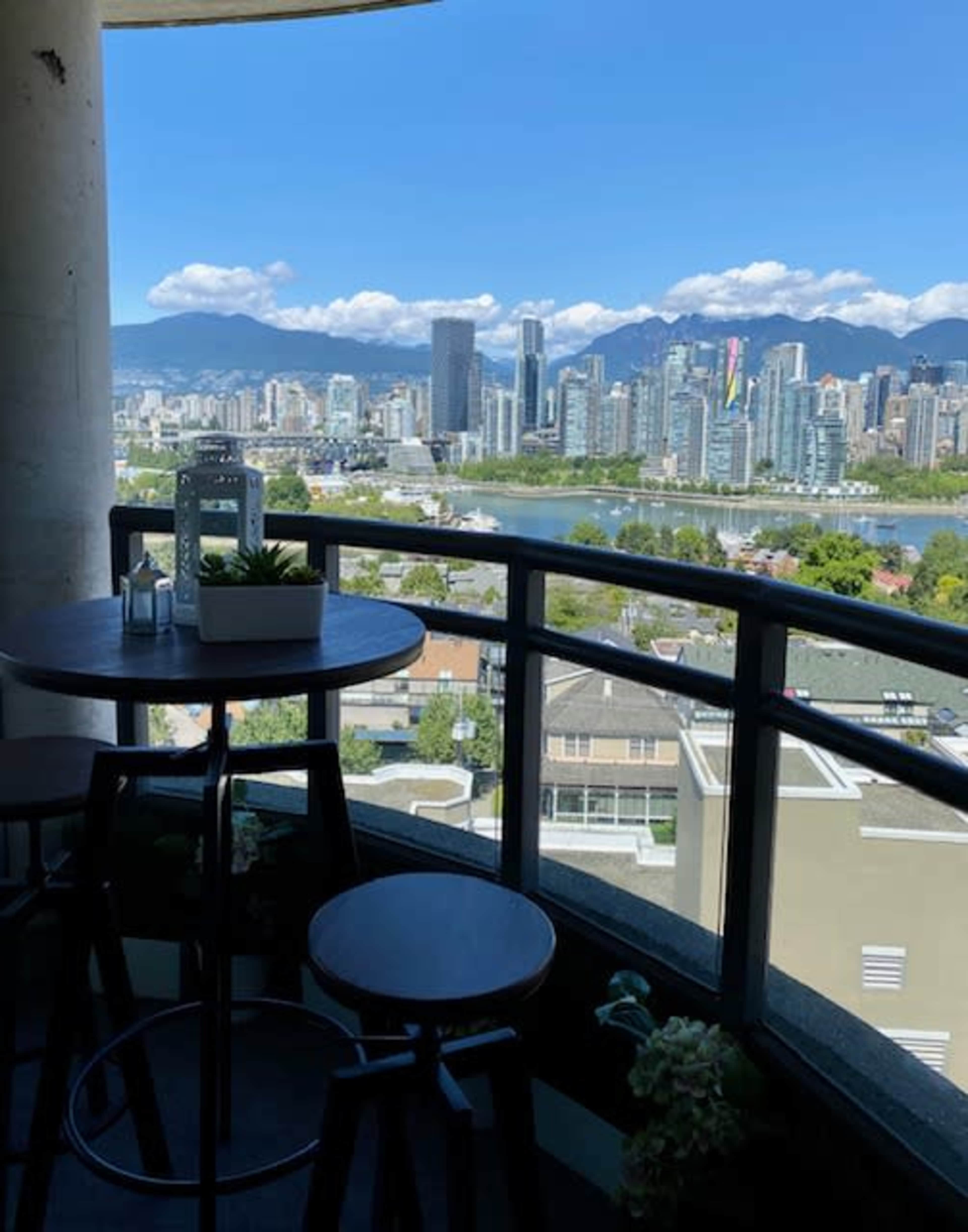 A balcony with a small table and two chairs overlooking a cityscape featuring high-rise buildings and mountains in the background.