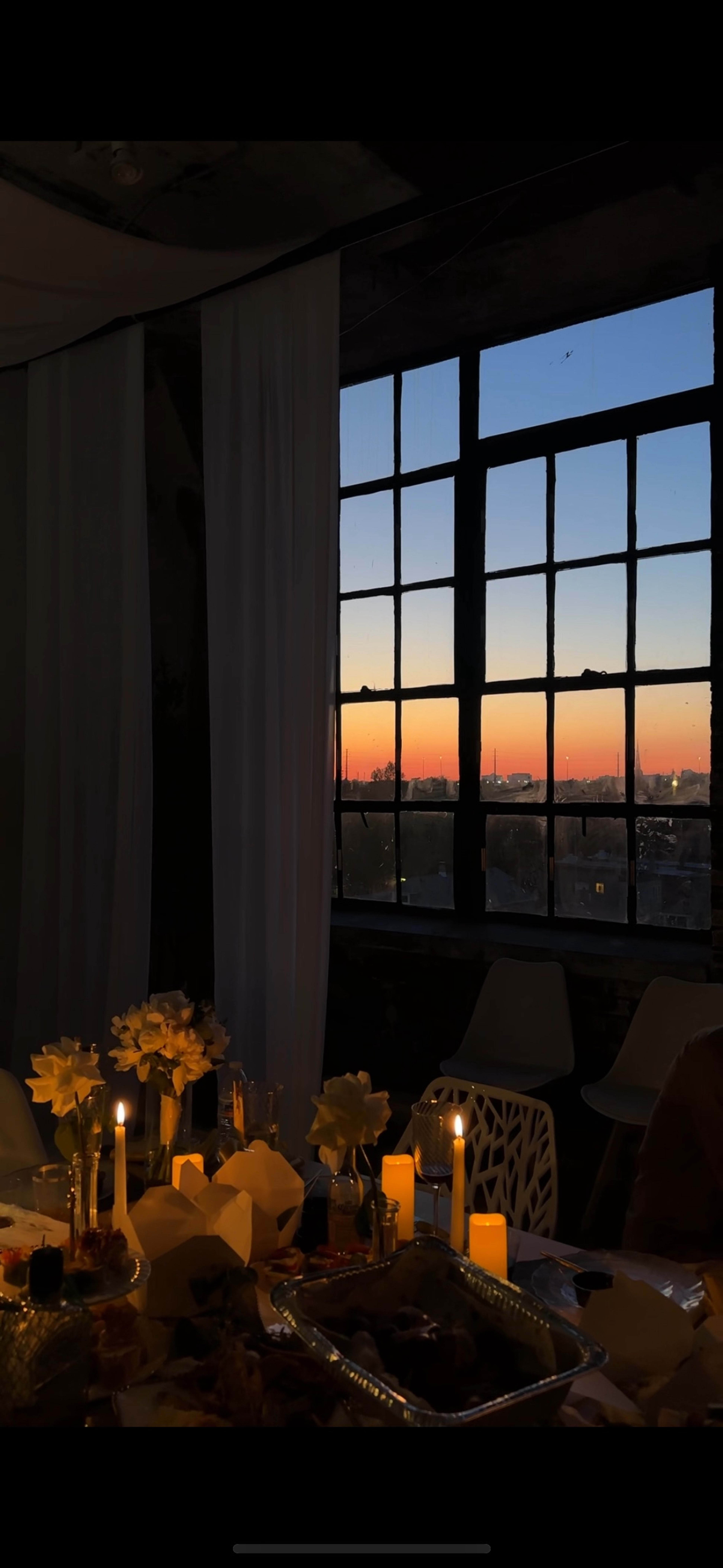 The image shows a table set with flowers and candles, framed by a large window displaying a sunset over a city skyline.
