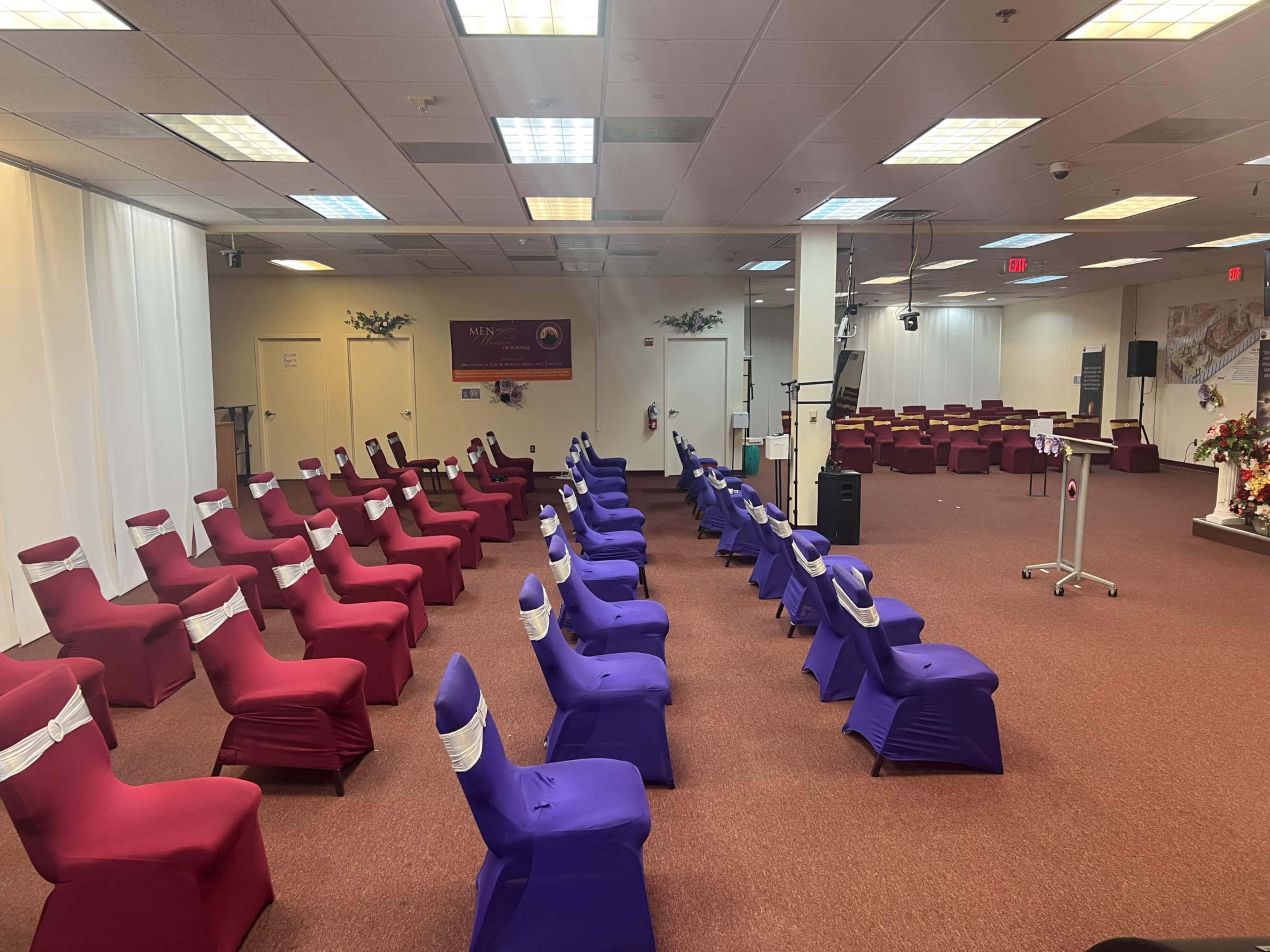 The image shows a hall set up for an event with rows of chairs covered in red and purple fabric.