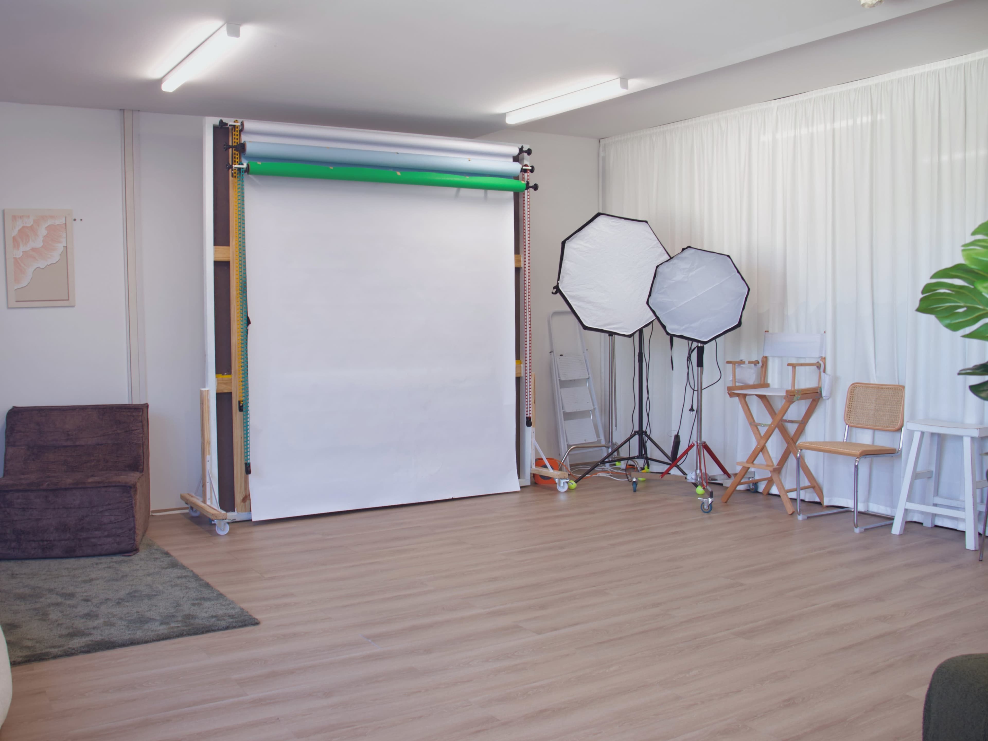 The image shows a photography studio with a blank backdrop, two light stands with soft boxes, a seating area, and a plant in the corner.