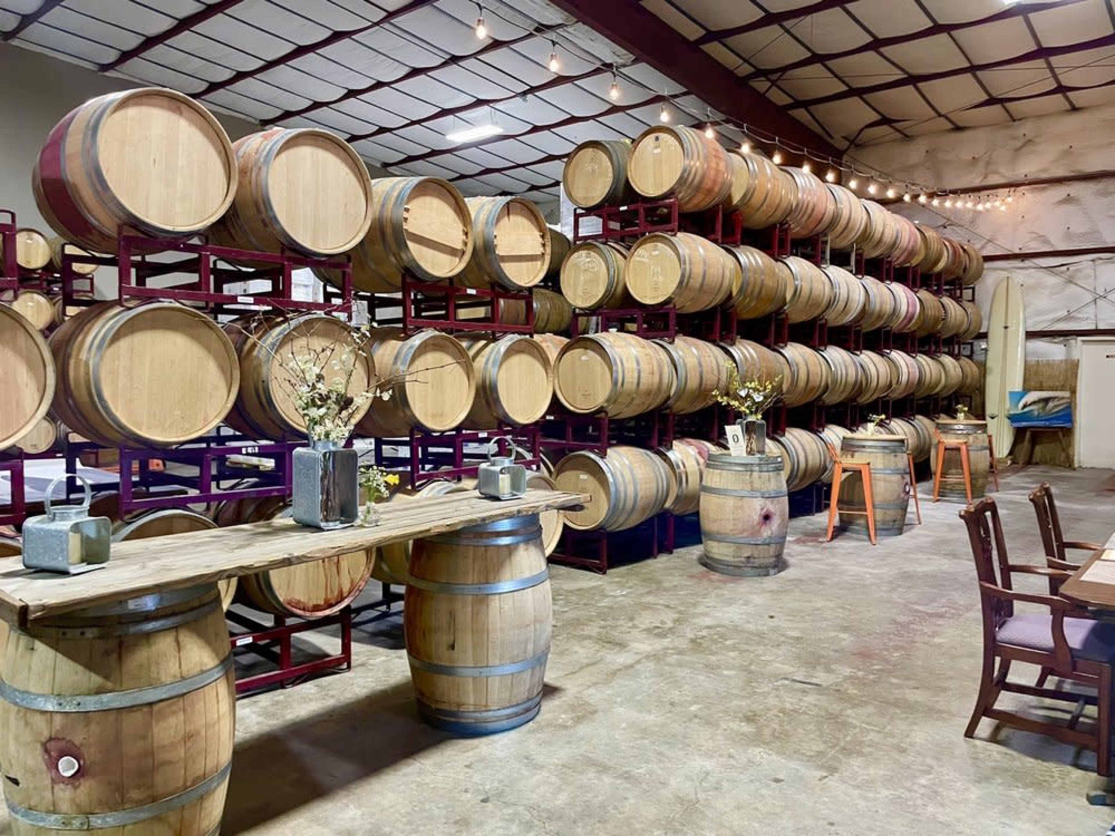 The image shows a winery interior with rows of stacked wooden barrels and a wooden table set with decor in front.