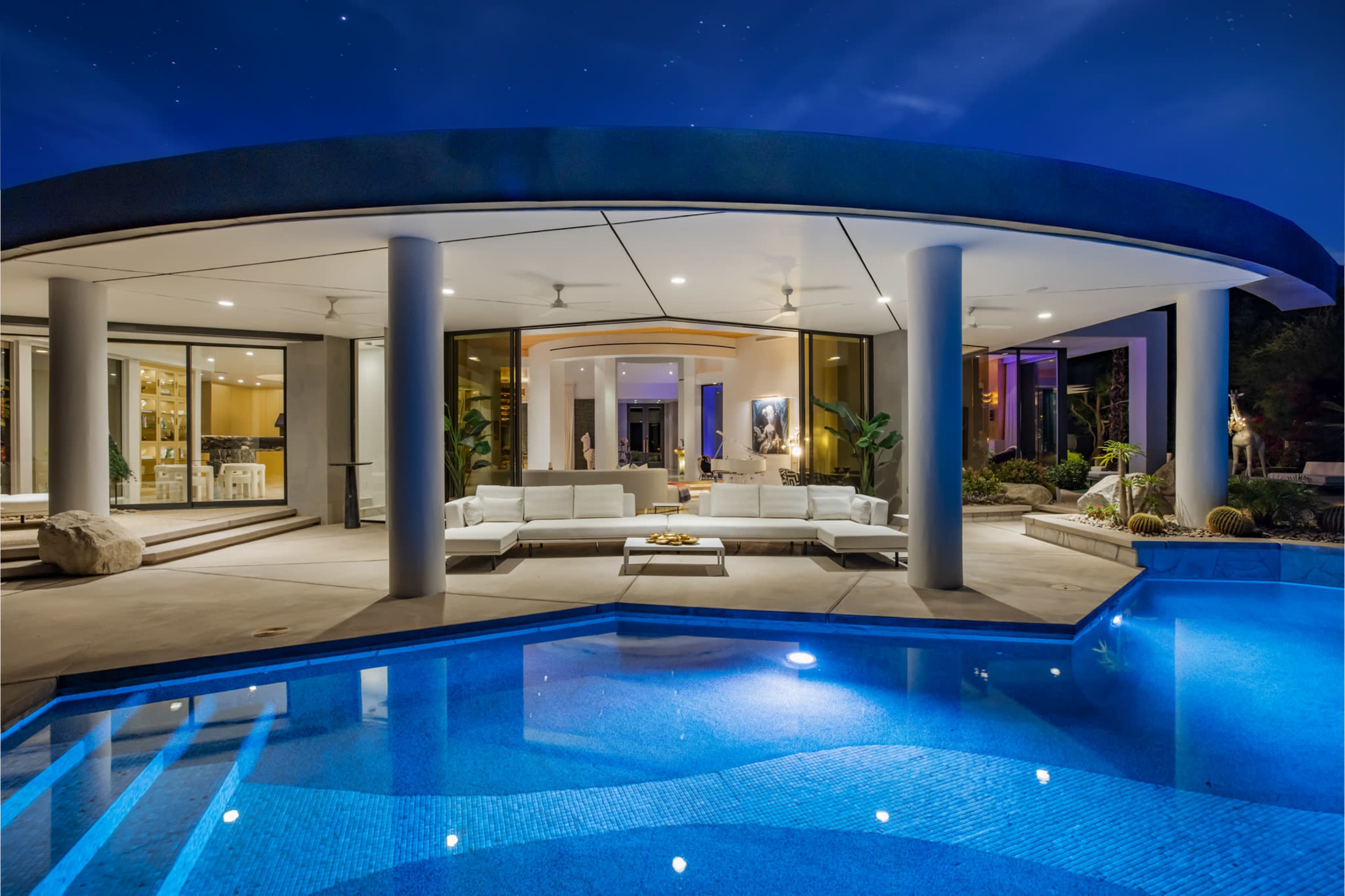 A modern outdoor lounge area is illuminated by soft lighting beside a swimming pool, with sleek white furniture and large glass windows showcasing the interior.