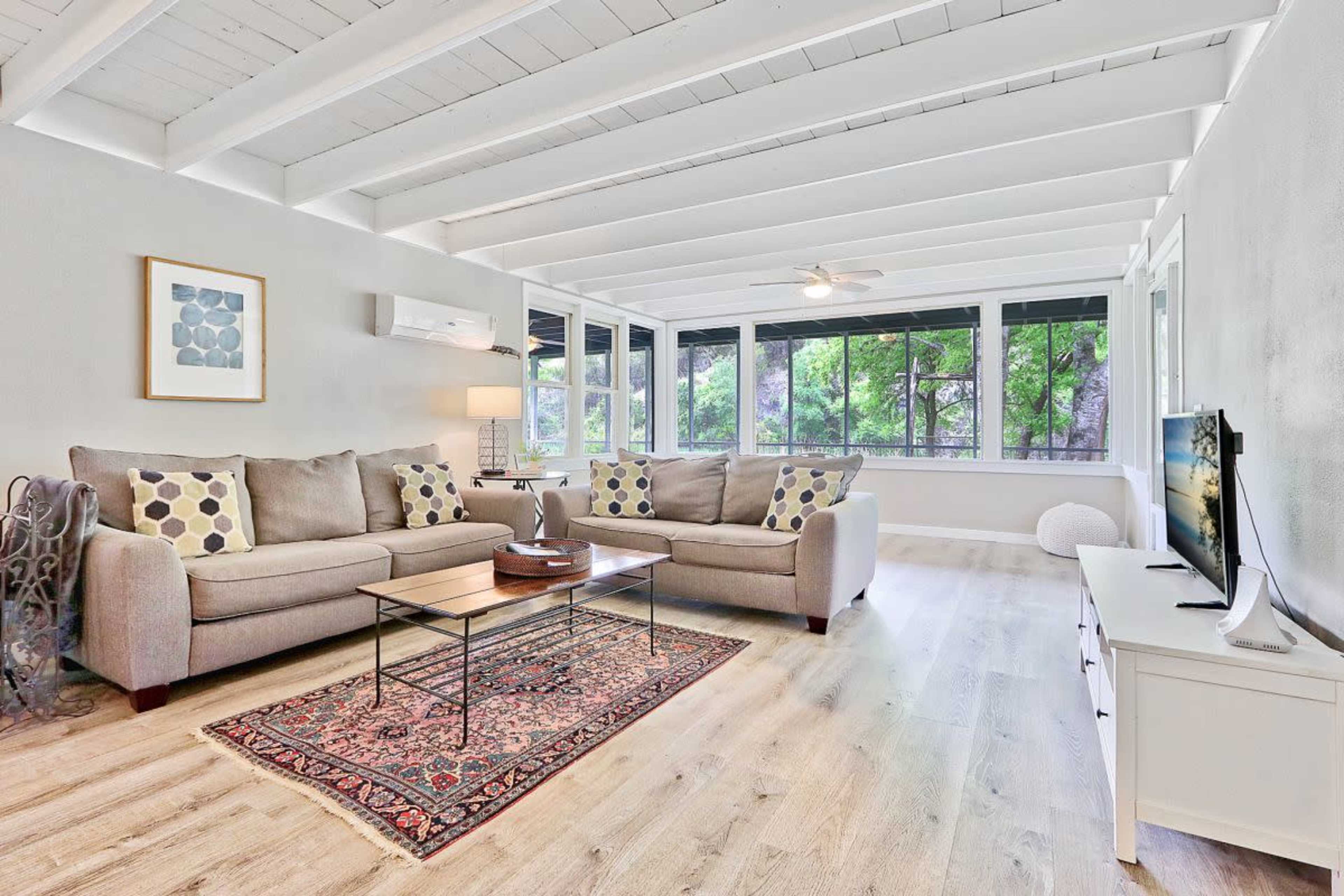 The image features a modern living room with two gray sofas, a coffee table, and a television, surrounded by large windows letting in natural light.