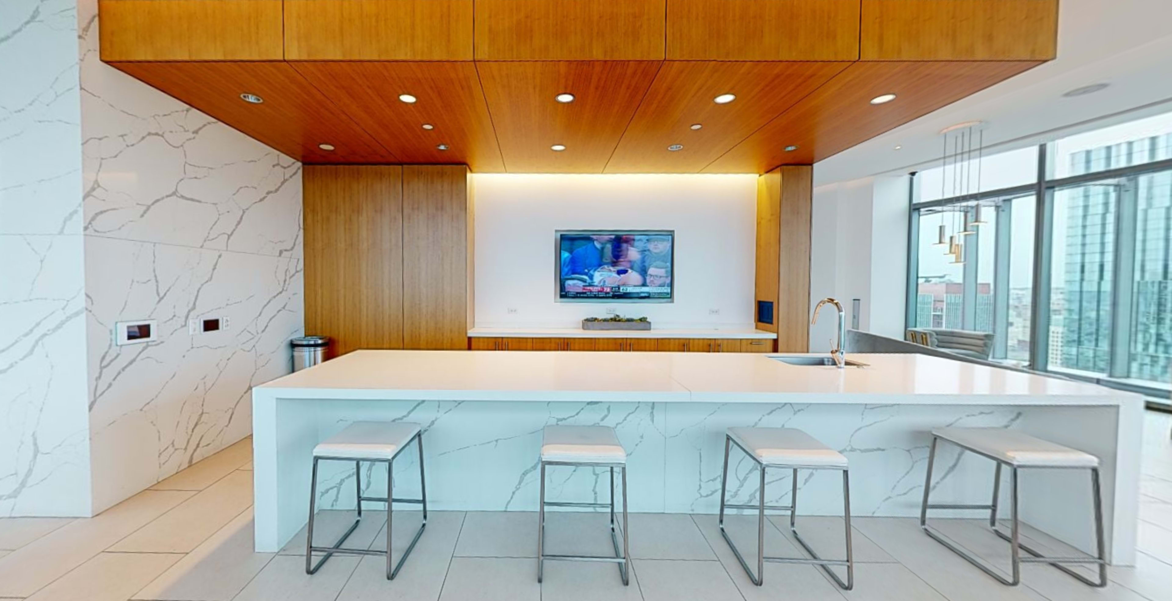 A modern kitchen with a large white island, four metal stools, wooden cabinetry, and a television mounted on the wall.
