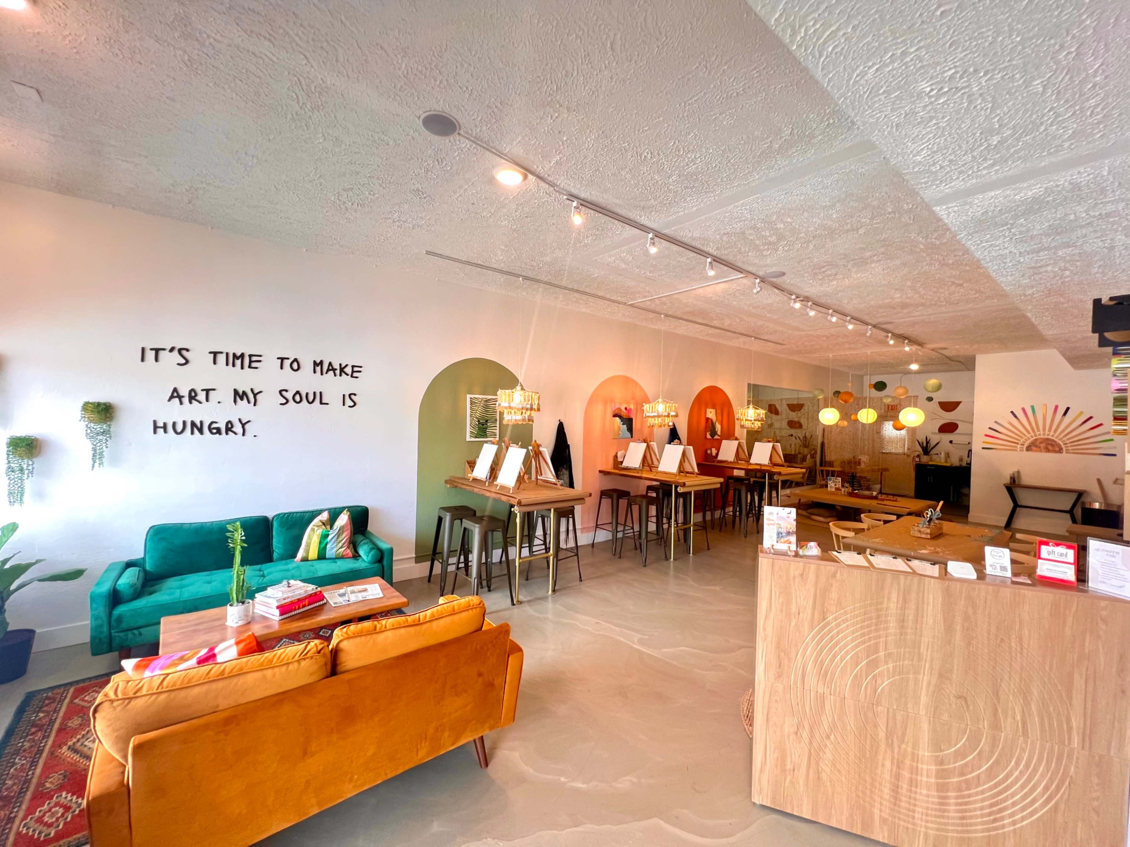 An interior space with a cozy seating area, a bar-like counter, and multiple tables set for art activities, featuring vibrant decor and text on the wall.
