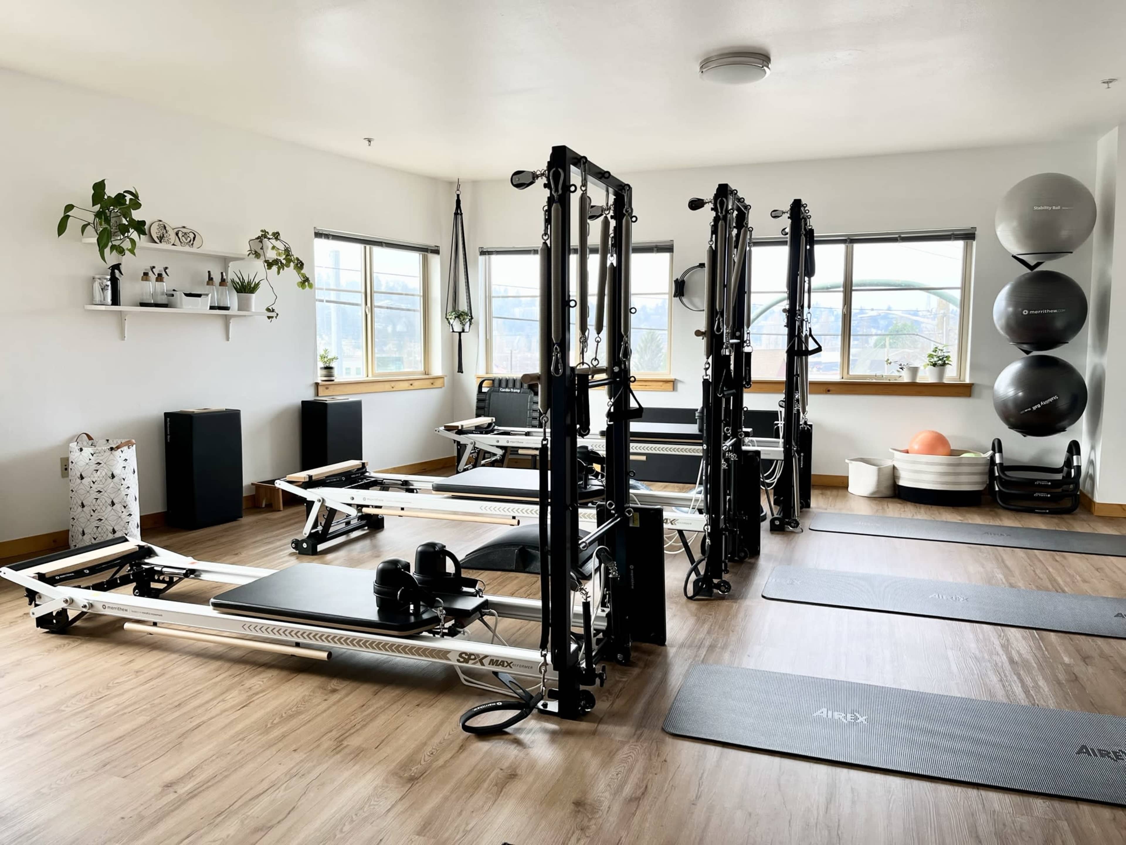 A well-lit fitness studio features reformer machines, exercise mats, and various workout equipment arranged neatly on a wooden floor.