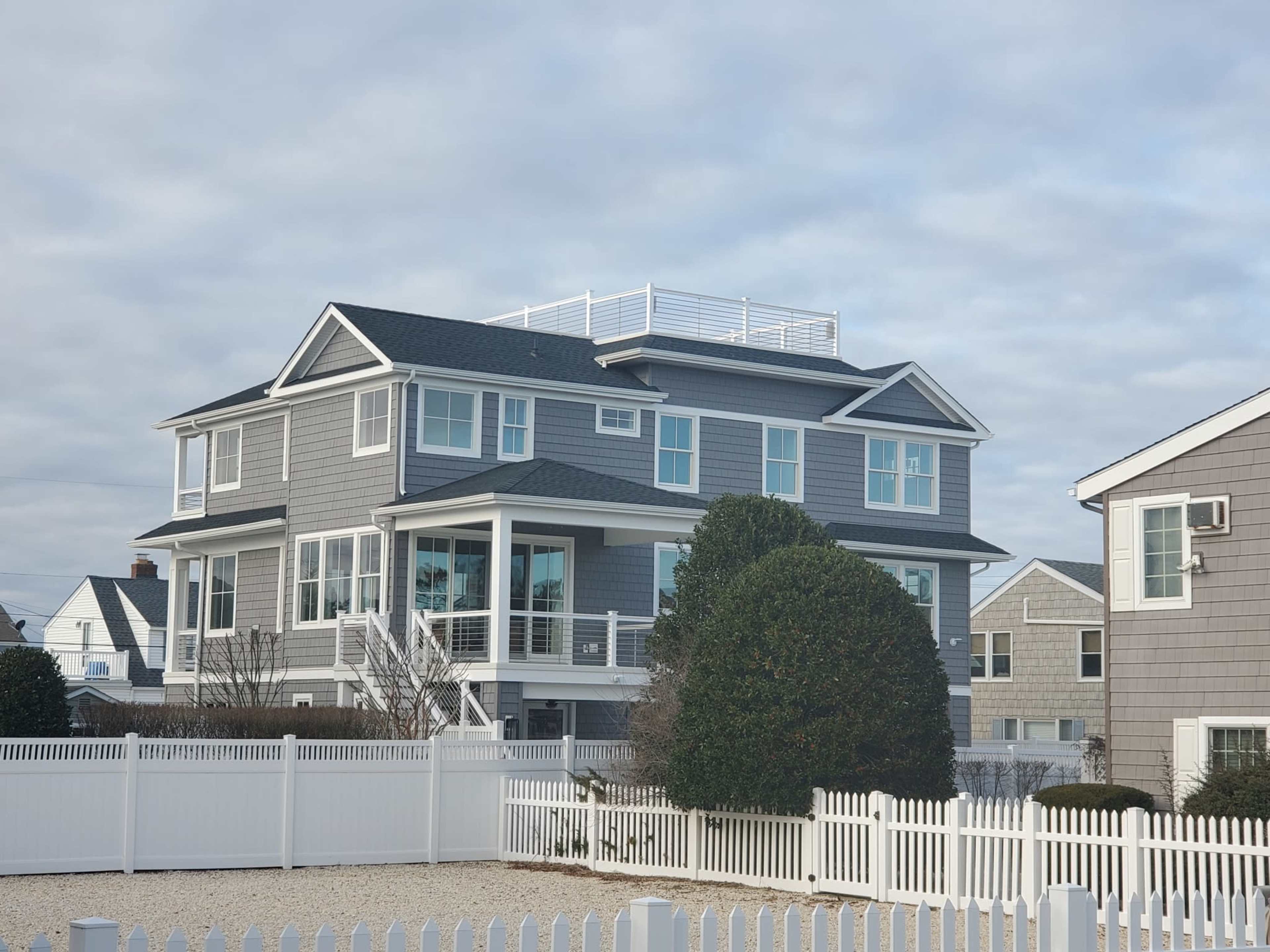 A large gray house with multiple balconies and a roof deck is surrounded by a white picket fence.
