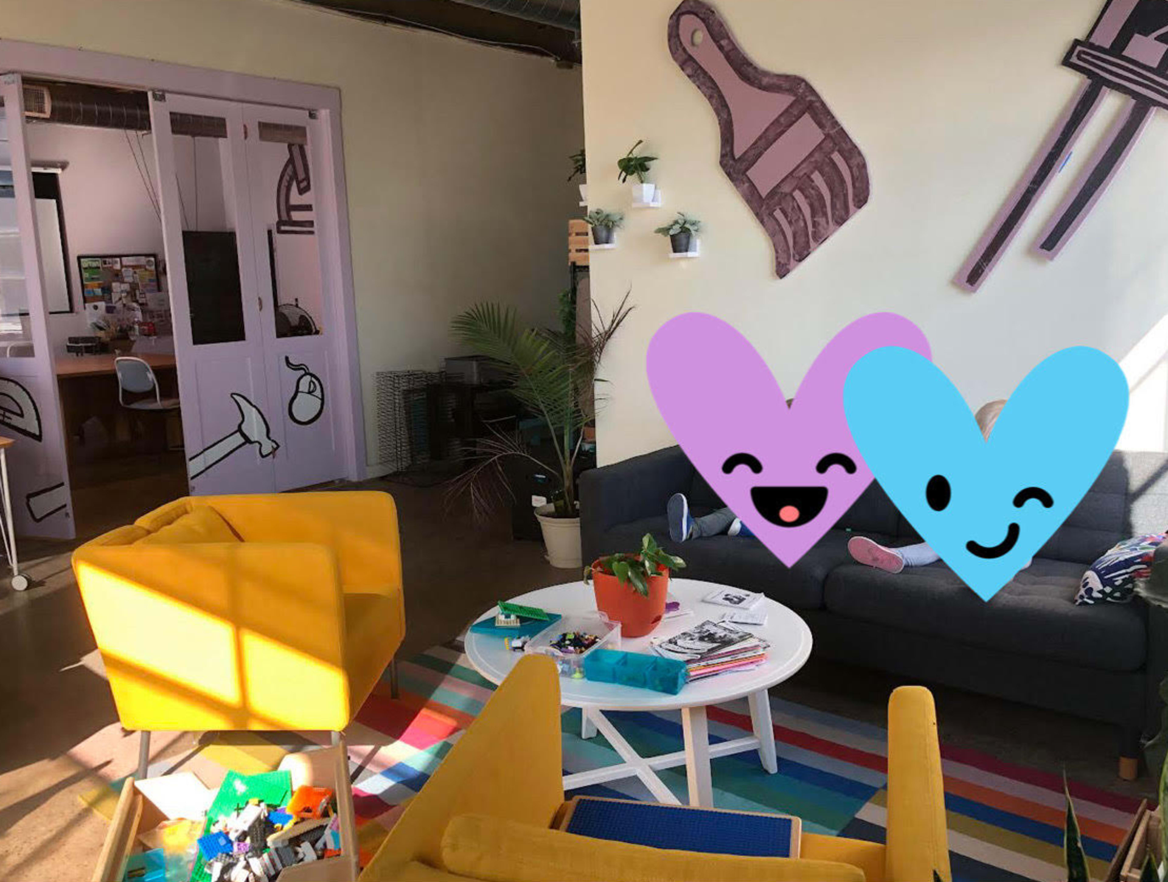 The image shows a brightly colored lounge area with a gray sofa, yellow chairs, a coffee table, and decorative plants, surrounded by playful wall art.