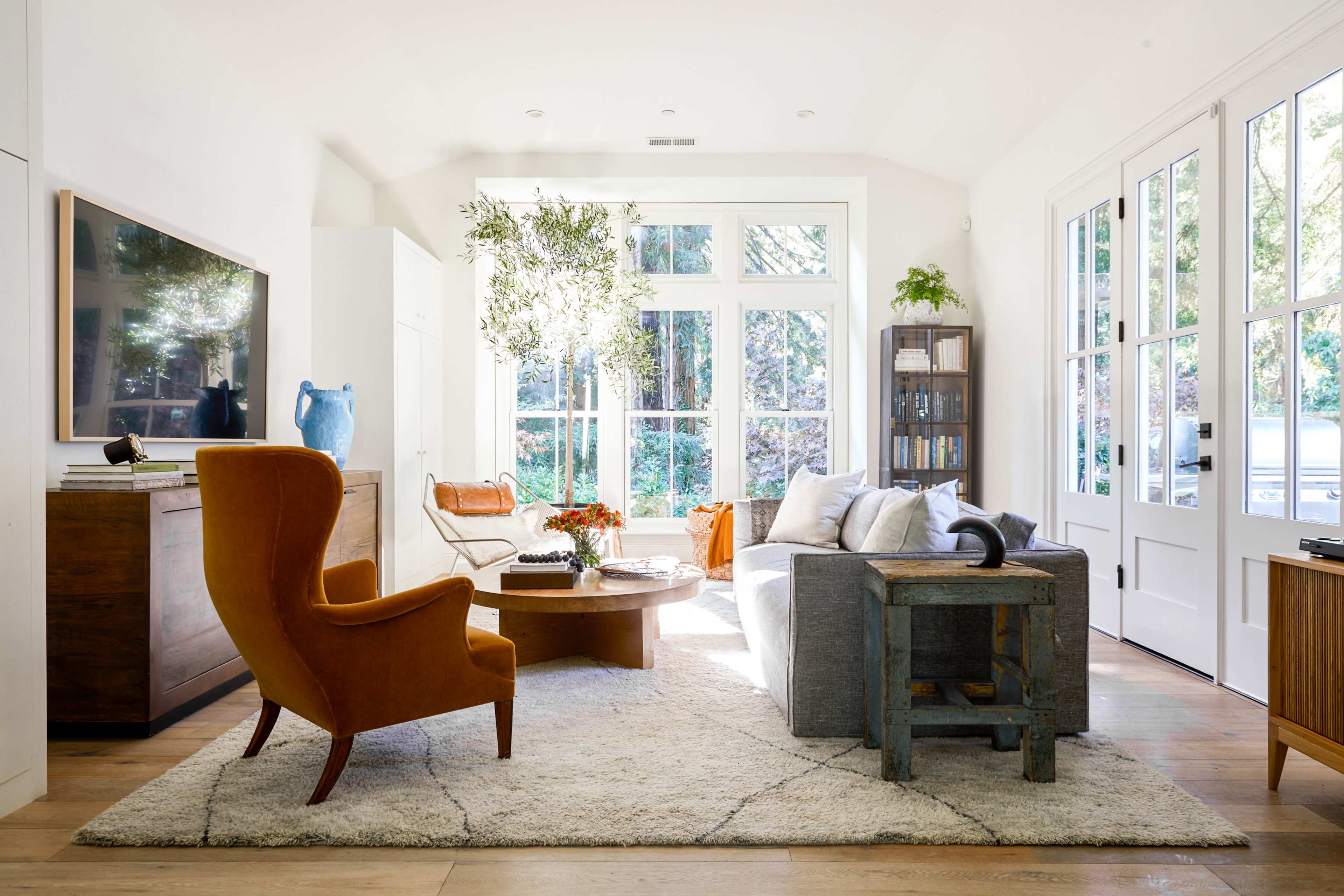 The living room features a mix of modern furniture, including a bright orange armchair, a gray sofa, and large windows that allow natural light to fill the space.