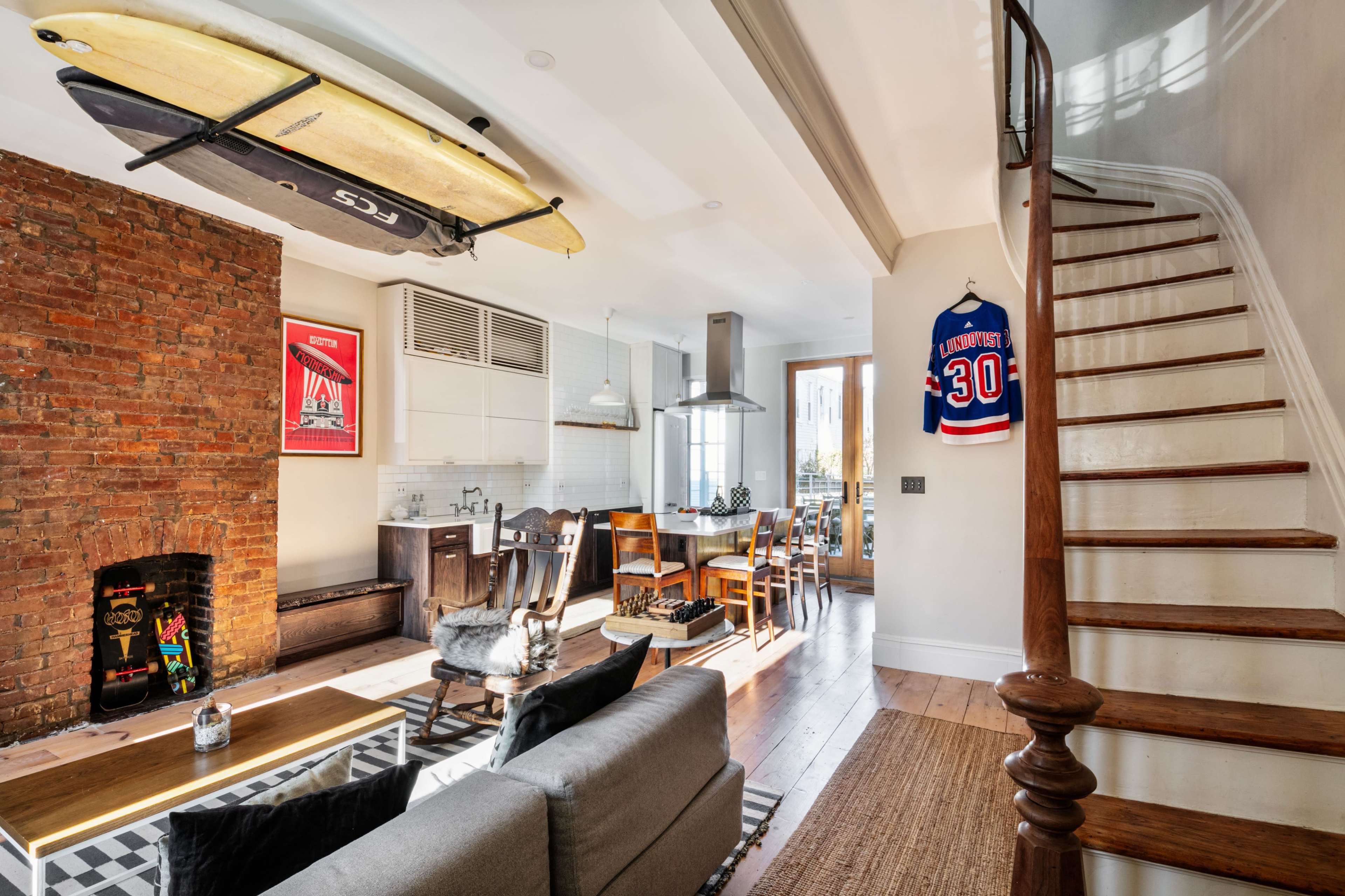 The image shows a modern living space featuring a brick wall, a wooden staircase, a surfboard mounted on the ceiling, and a dining area with a large table.