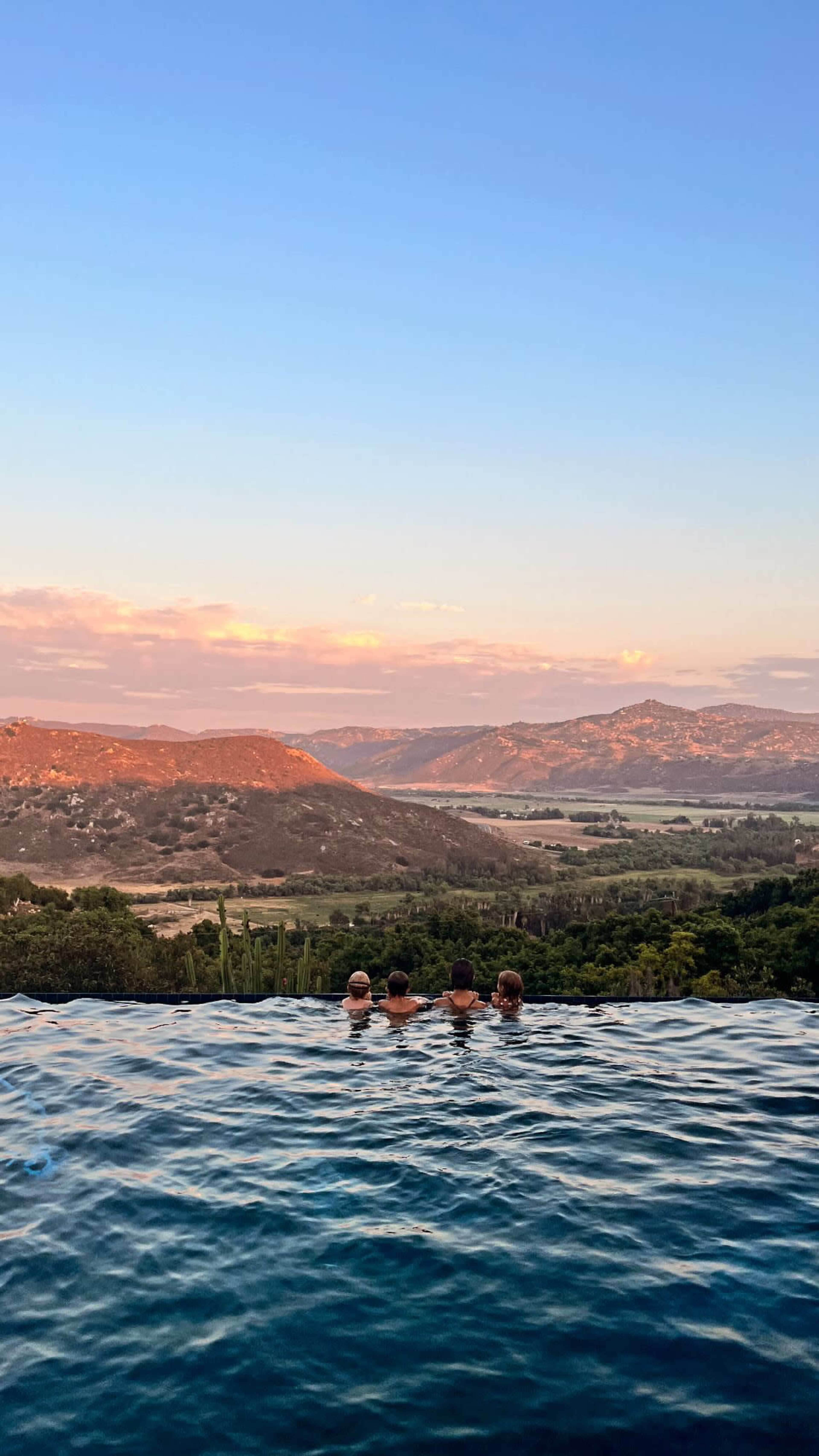 Three people sit at the edge of an infinity pool, overlooking a scenic landscape of rolling hills and a sunset sky.