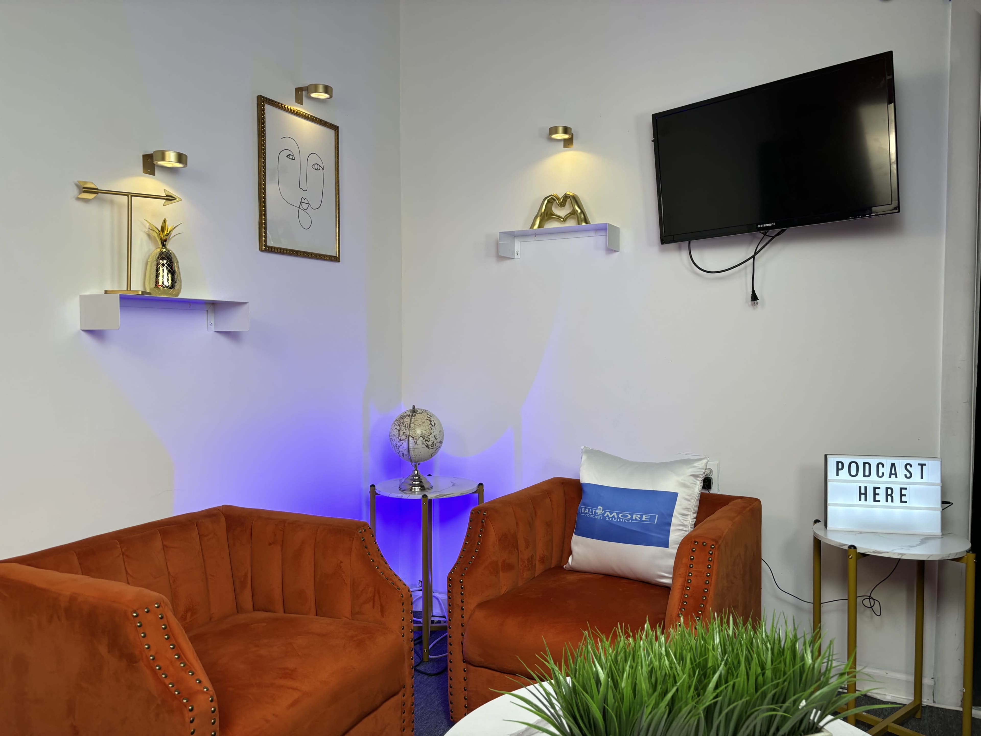 The image depicts a cozy studio space with two orange armchairs, a small round table, a television mounted on the wall, and decorative elements including a globe and a lightbox that says "PODCAST HERE."