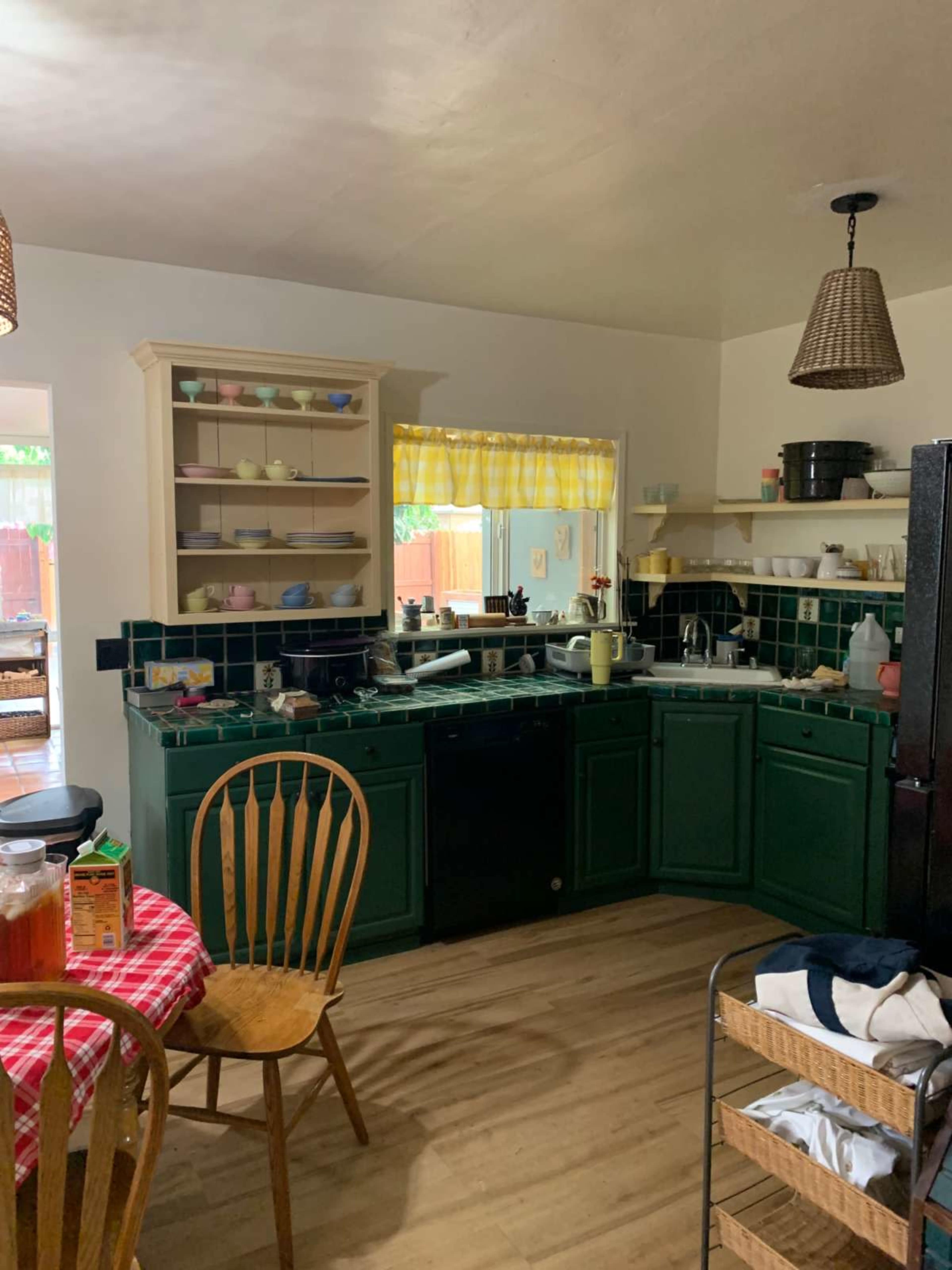 The image shows a kitchen with green cabinets, a round table covered in a red checkered tablecloth, wooden chairs, and a variety of dishes displayed on shelves.