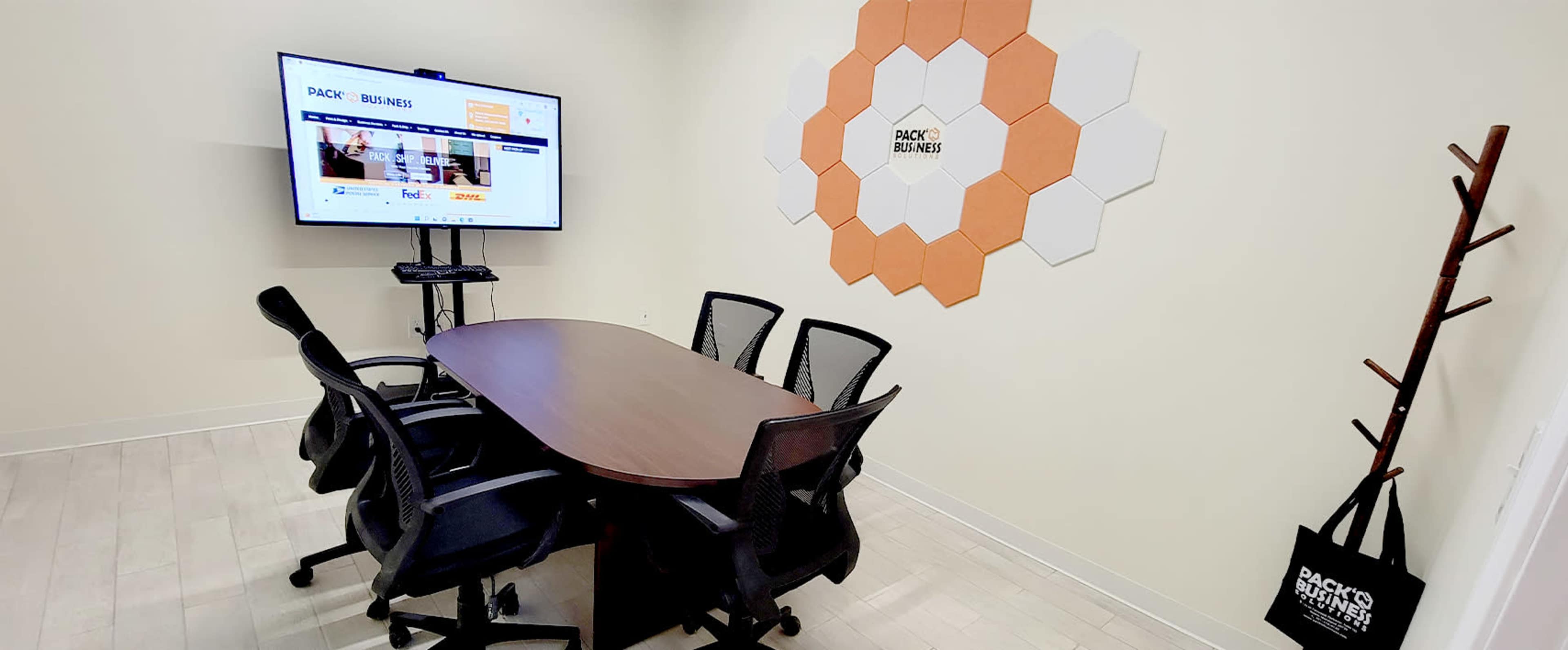 A meeting room with a rectangular wooden table, six black mesh chairs, a large screen displaying a website, and a wall decorated with hexagonal orange and white panels.