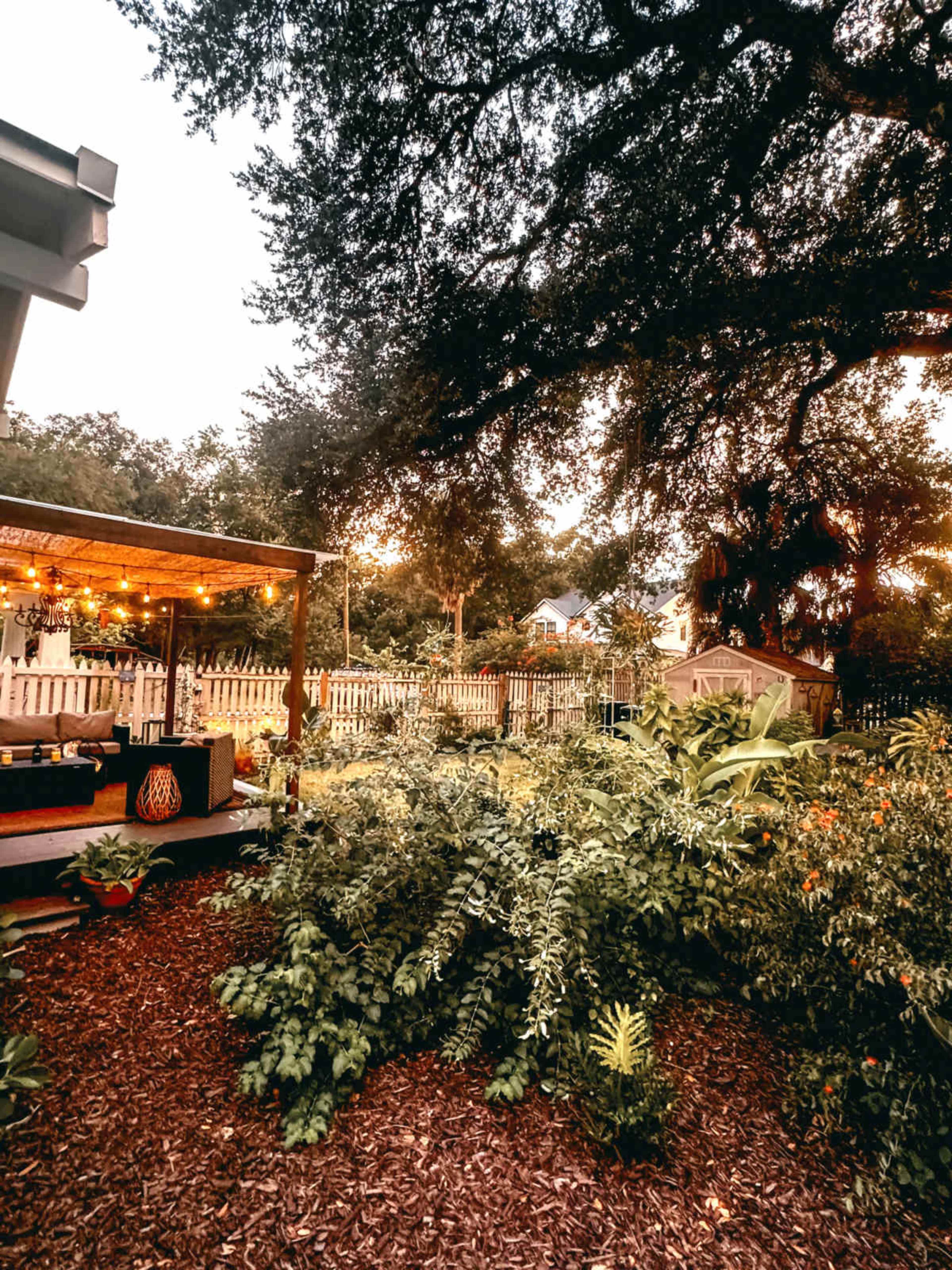 The image shows a backyard garden with lush greenery, decorative lighting, and a wooden deck surrounded by a wooden fence.