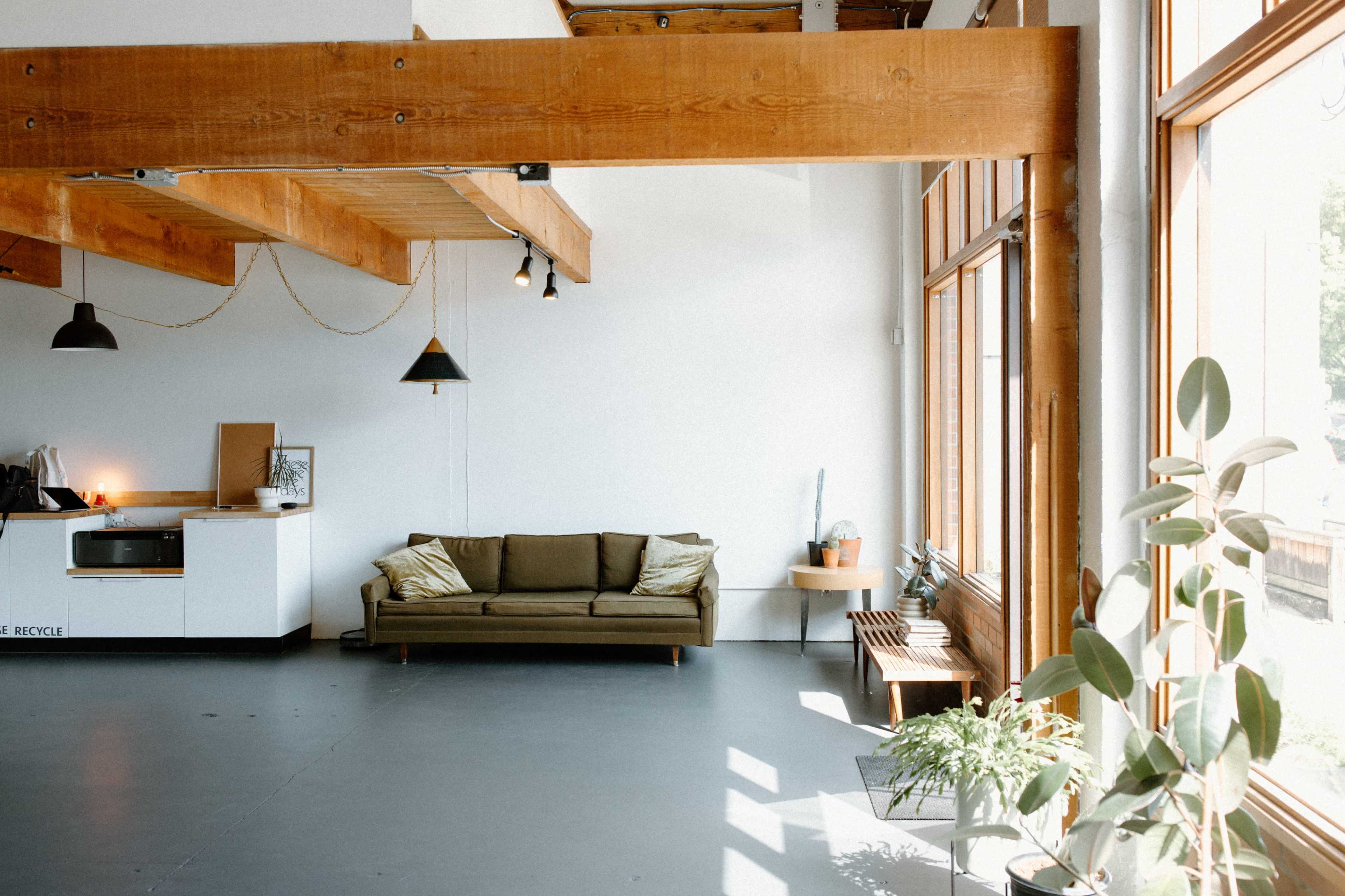 A spacious interior with wooden beams, large windows, a green sofa, a kitchen area, and various plants.