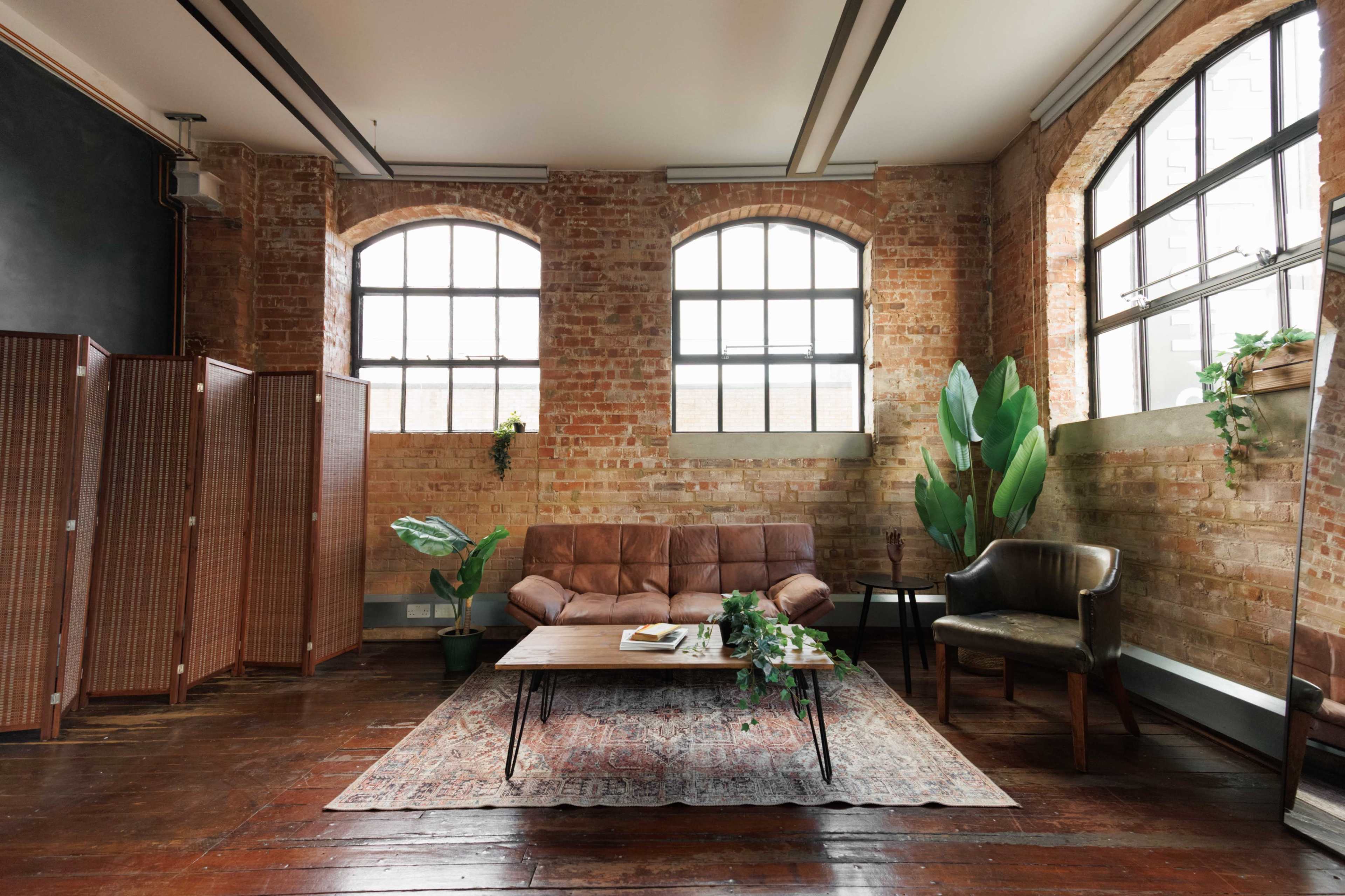 The image shows a cozy living area with a brown leather sofa, a dark wooden coffee table, and large windows letting in natural light, alongside green plants and a patterned rug.