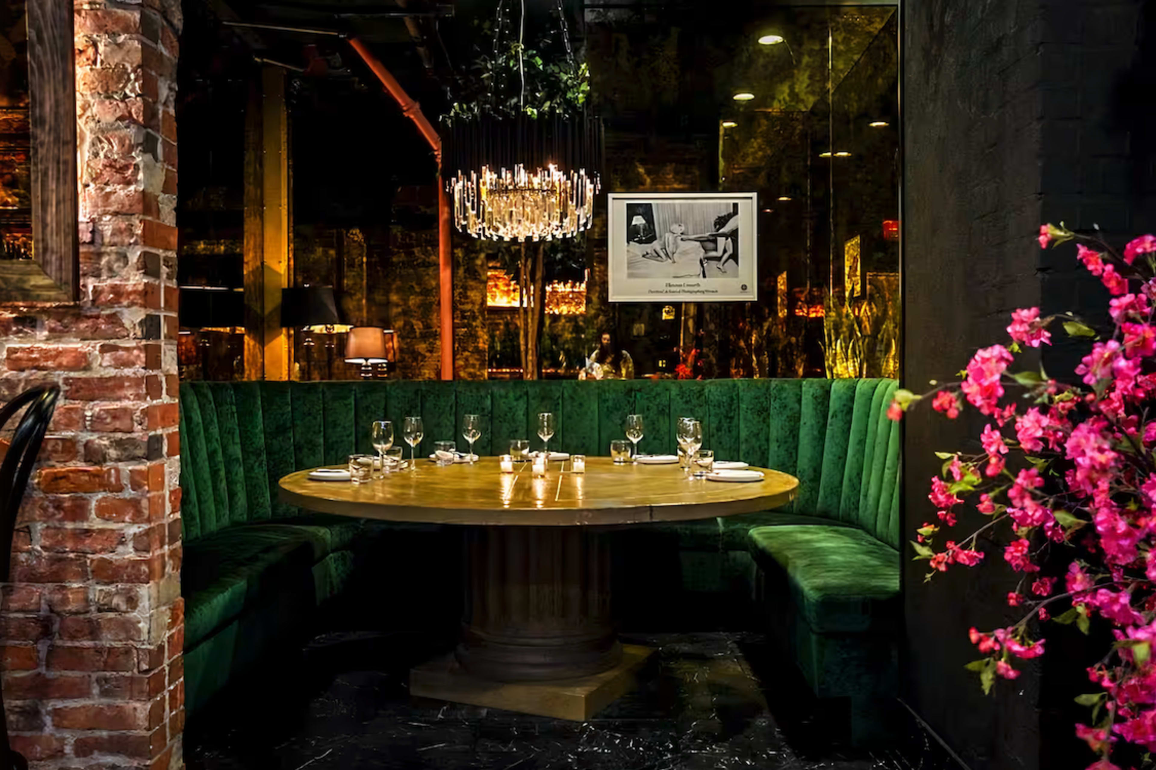 A round wooden table with place settings is centered in a green upholstered booth, surrounded by exposed brick walls and decorative elements like a chandelier and flowers.